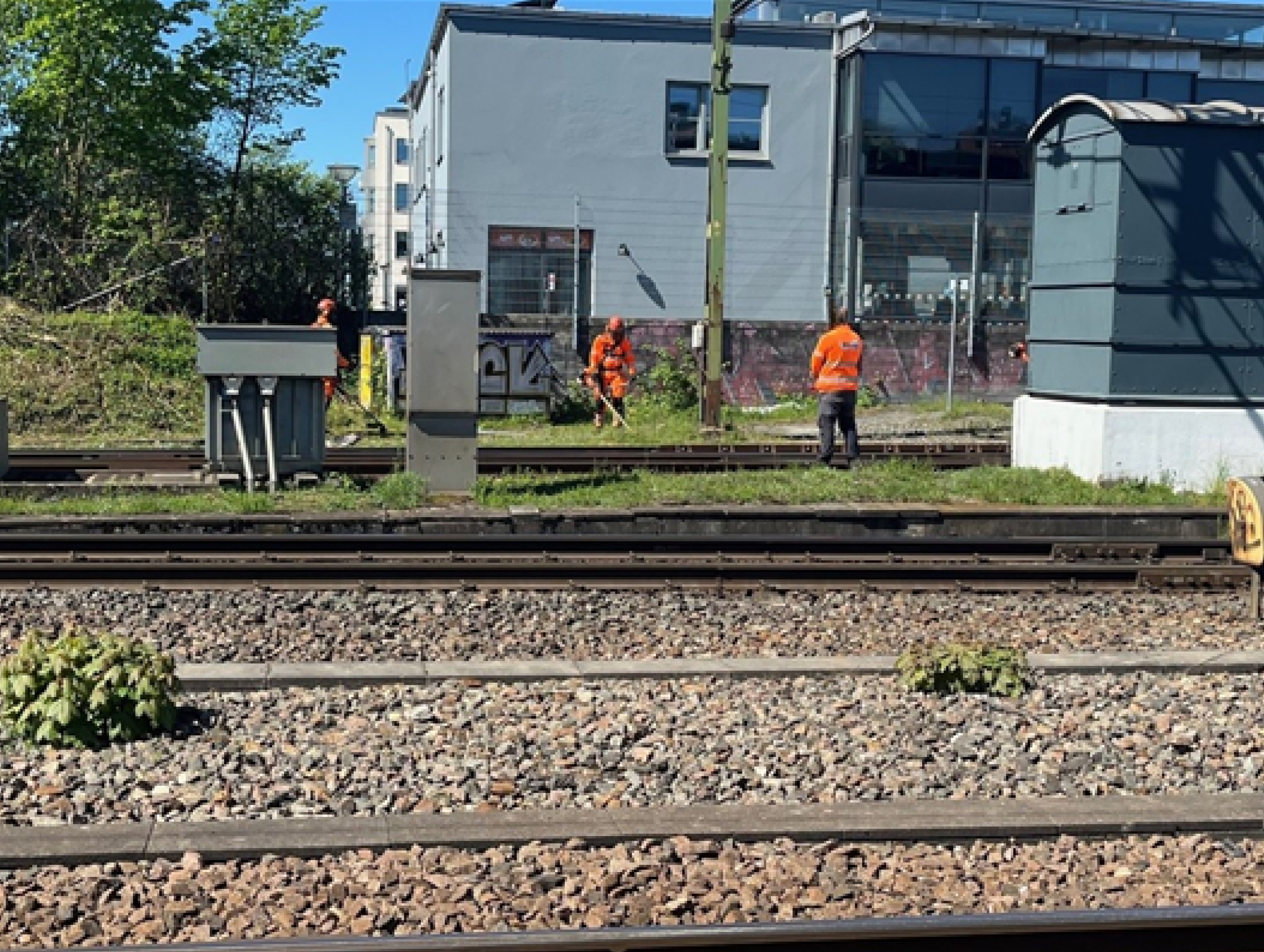 Fyra arbetare i orange skyddskläder arbetar vid järnvägsspår bredvid en grå byggnad under dagen.