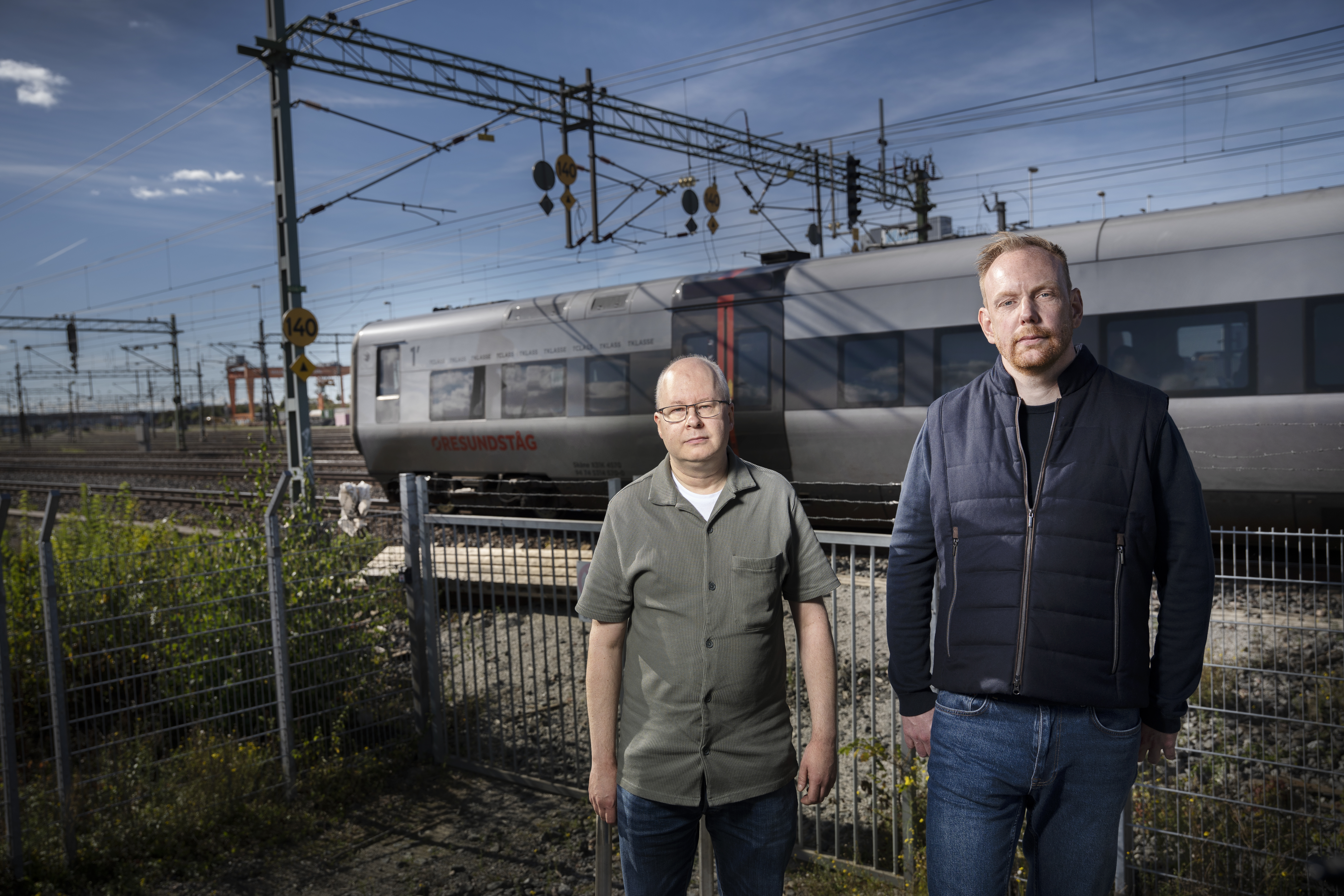Janne Petersson och Martin Nilsson står framför ett metallstängsel. I bakgrunden syns ett tåg och järnvägsspår under en himmel.