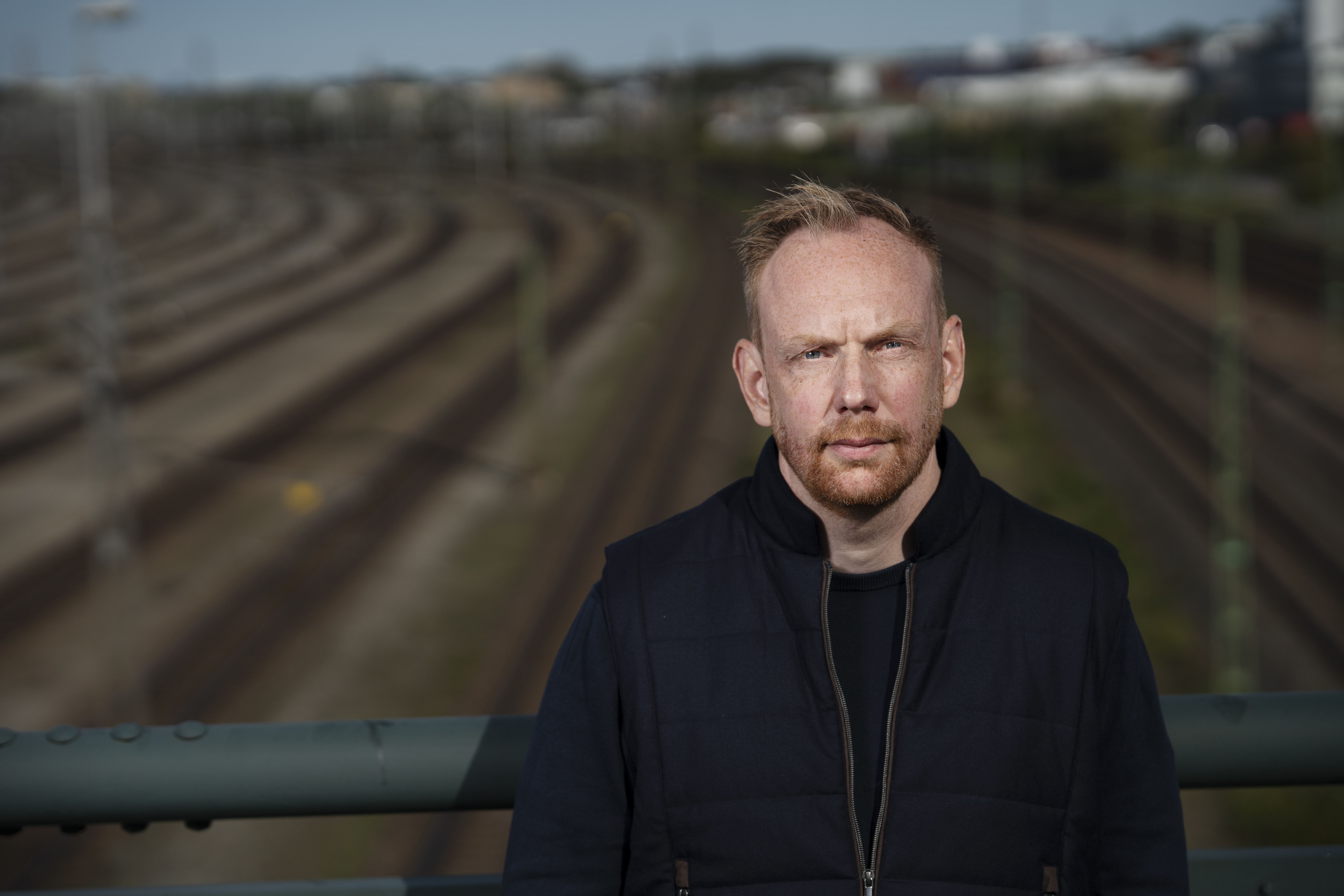 En man med ljus hud och kort rött hår står på en bro och tittar ut över flera järnvägsspår.