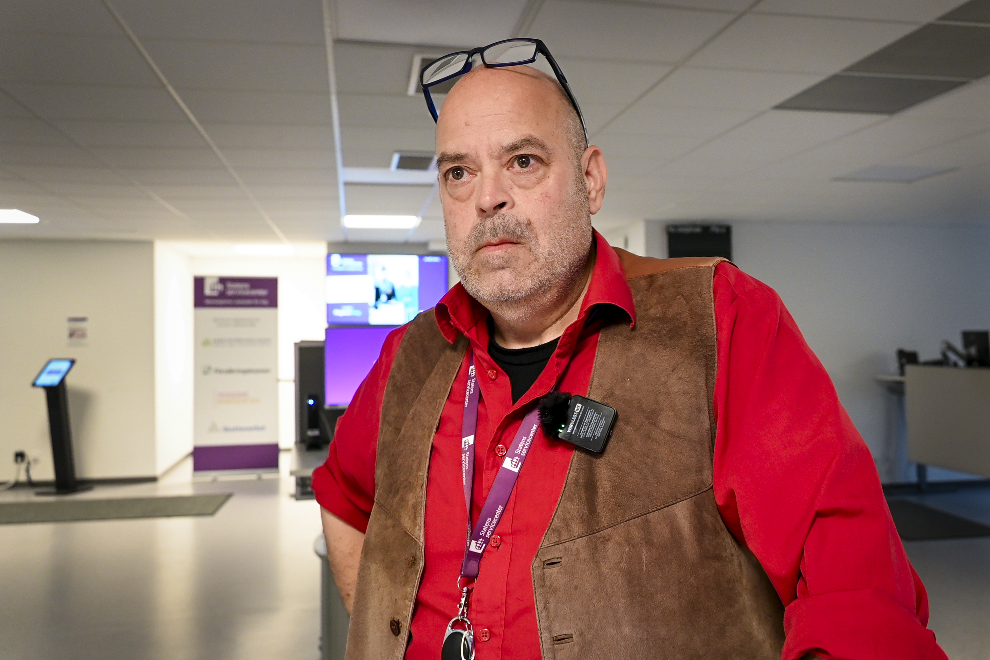 Peter Hjelm med röd skjorta, brun väst och glasögon står i ett kontor med en namnbricka och elektroniskt passerkort runt halsen.
