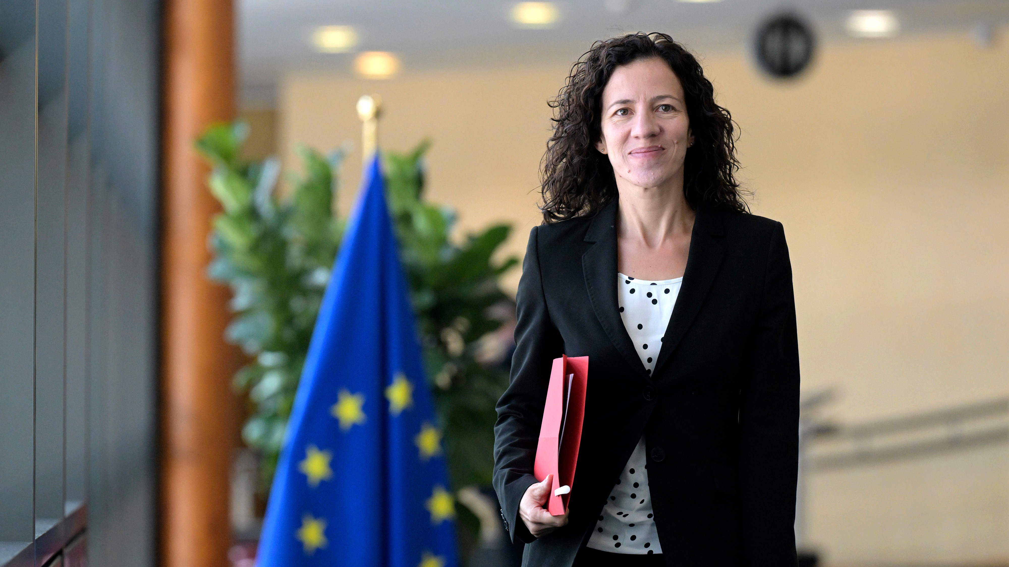 En kvinna i svart kostym håller en röd mapp och står inomhus nära en EU-flagga, med växter och ljus i bakgrunden.