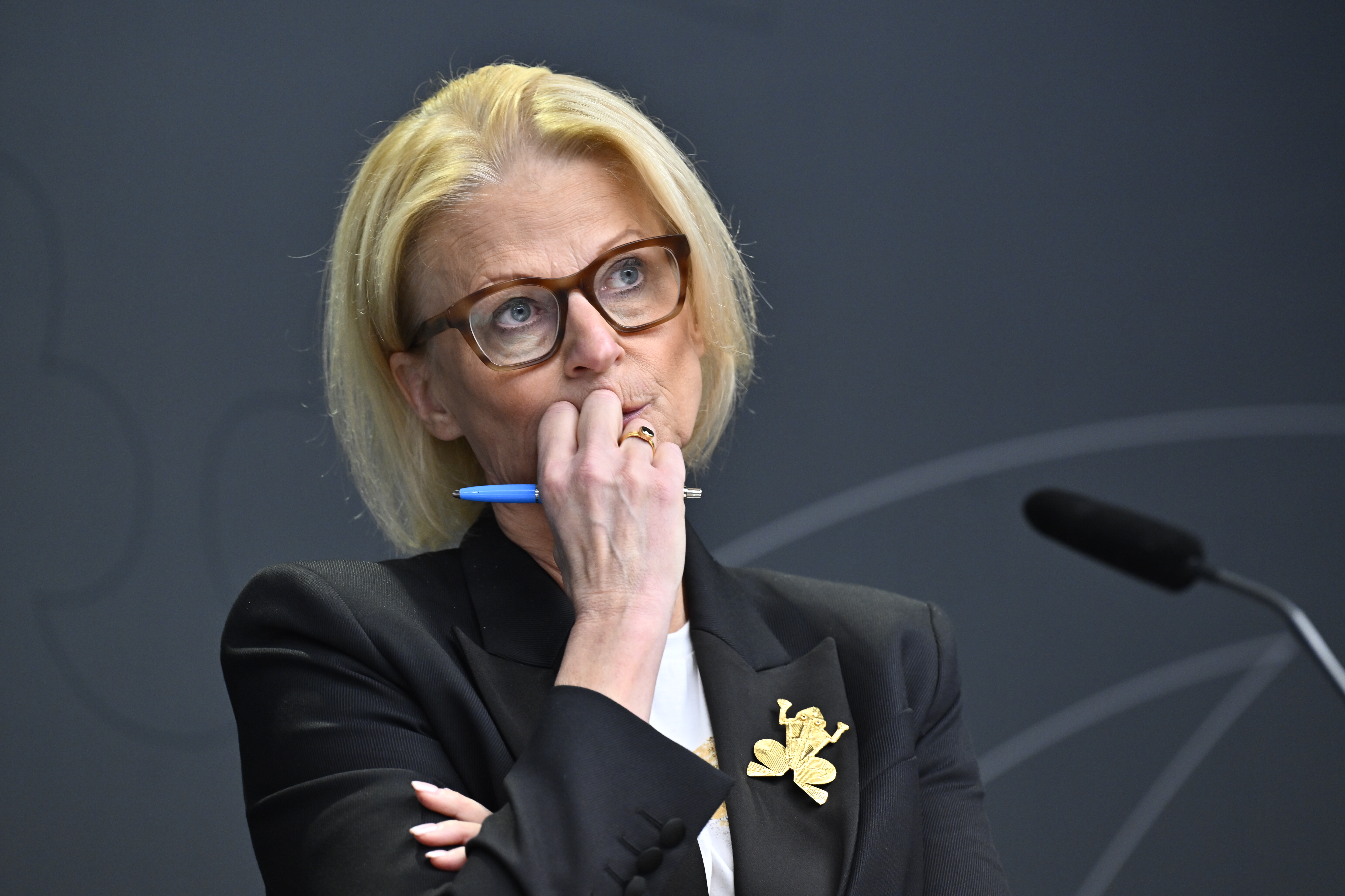 En kvinna med blont hår och glasögon, klädd i svart kavaj och hållande en penna, står framför en mikrofon.