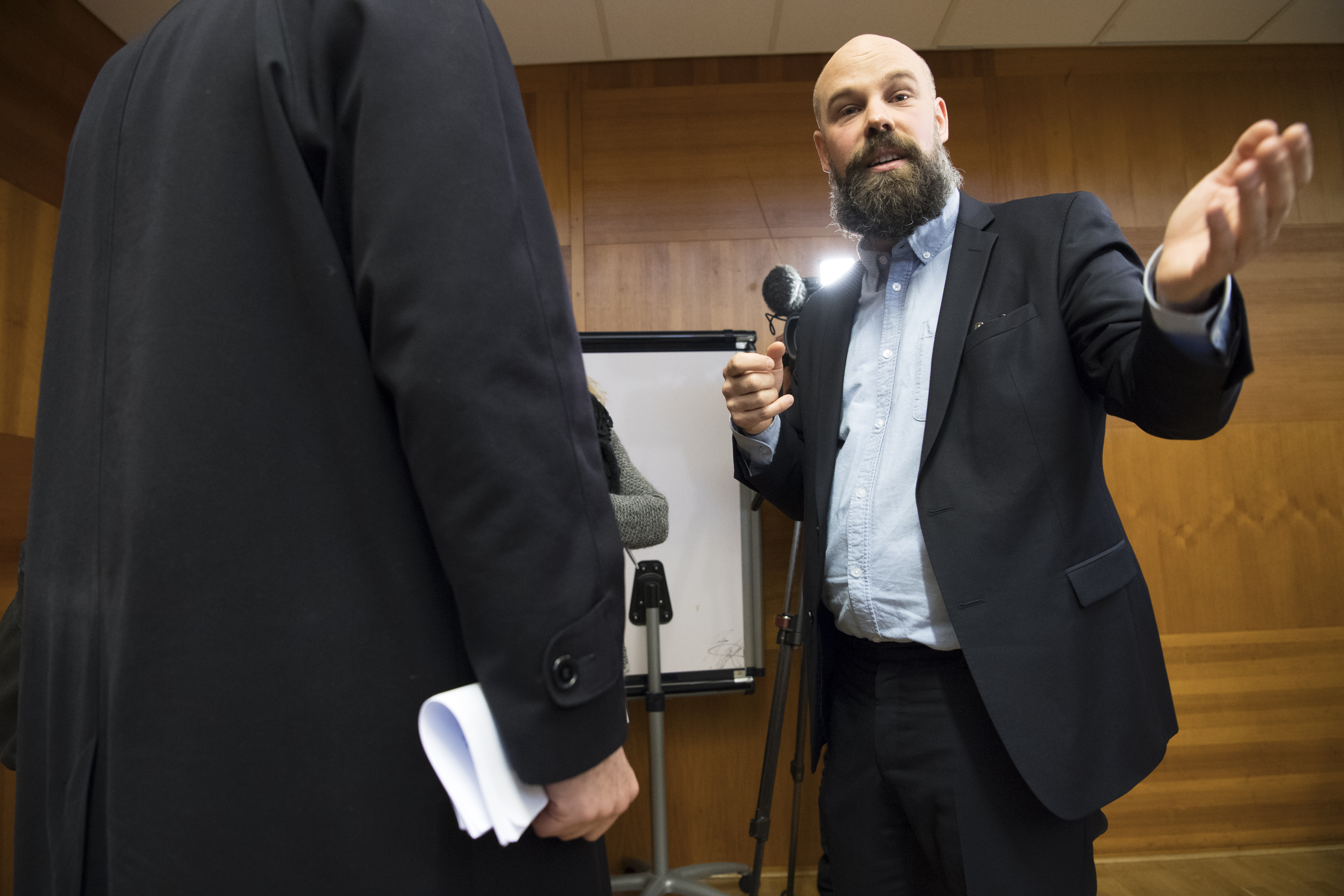 En man med mikrofon gestikulerar medan han talar med en annan person i kostym framför ett blädderblock i ett trärum.