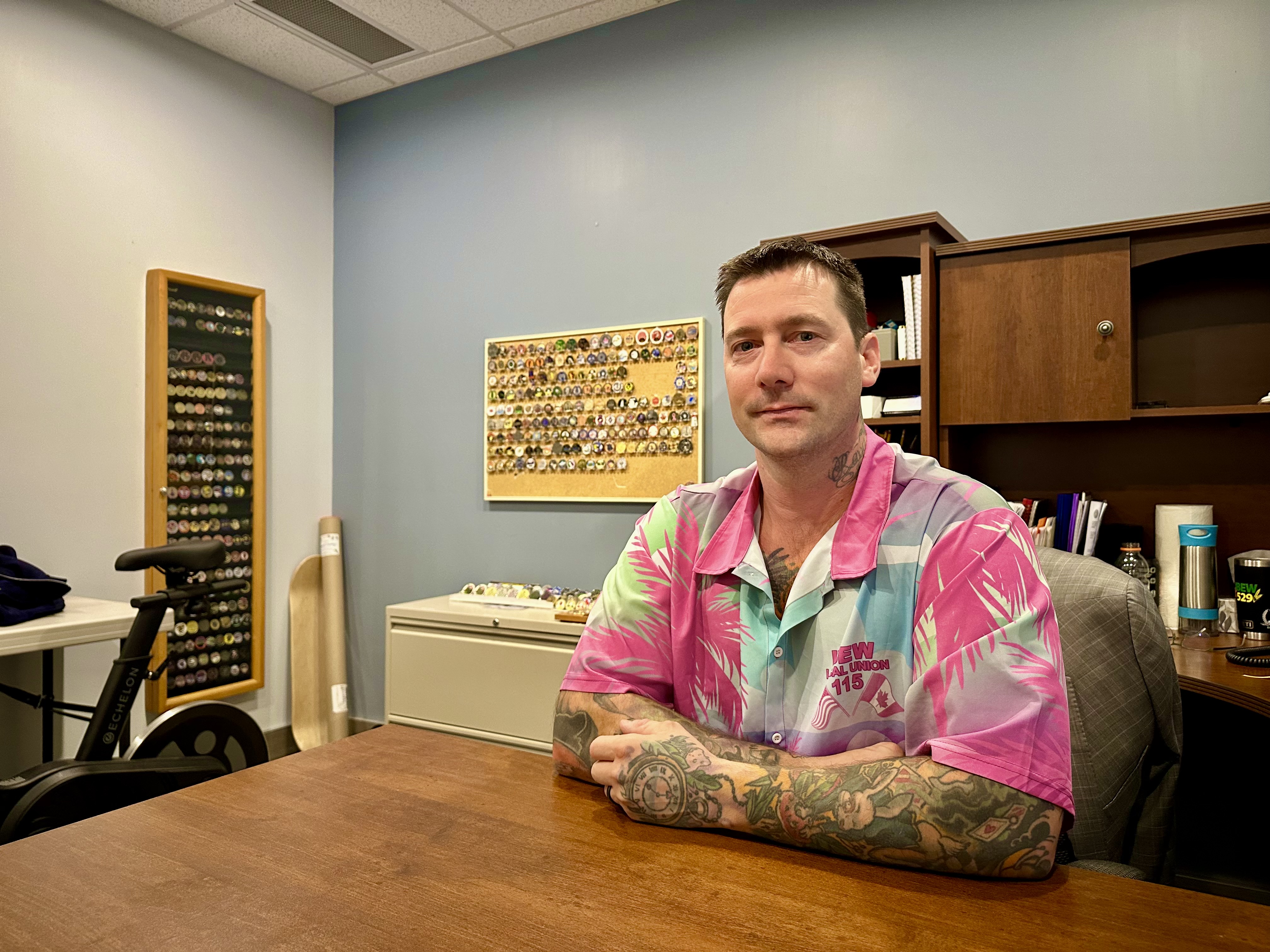 En man med tatuerade armar sitter vid ett skrivbord i ett kontor och har på sig en rosa och blå skjorta. Mynt eller märken syns på väggen bakom honom.