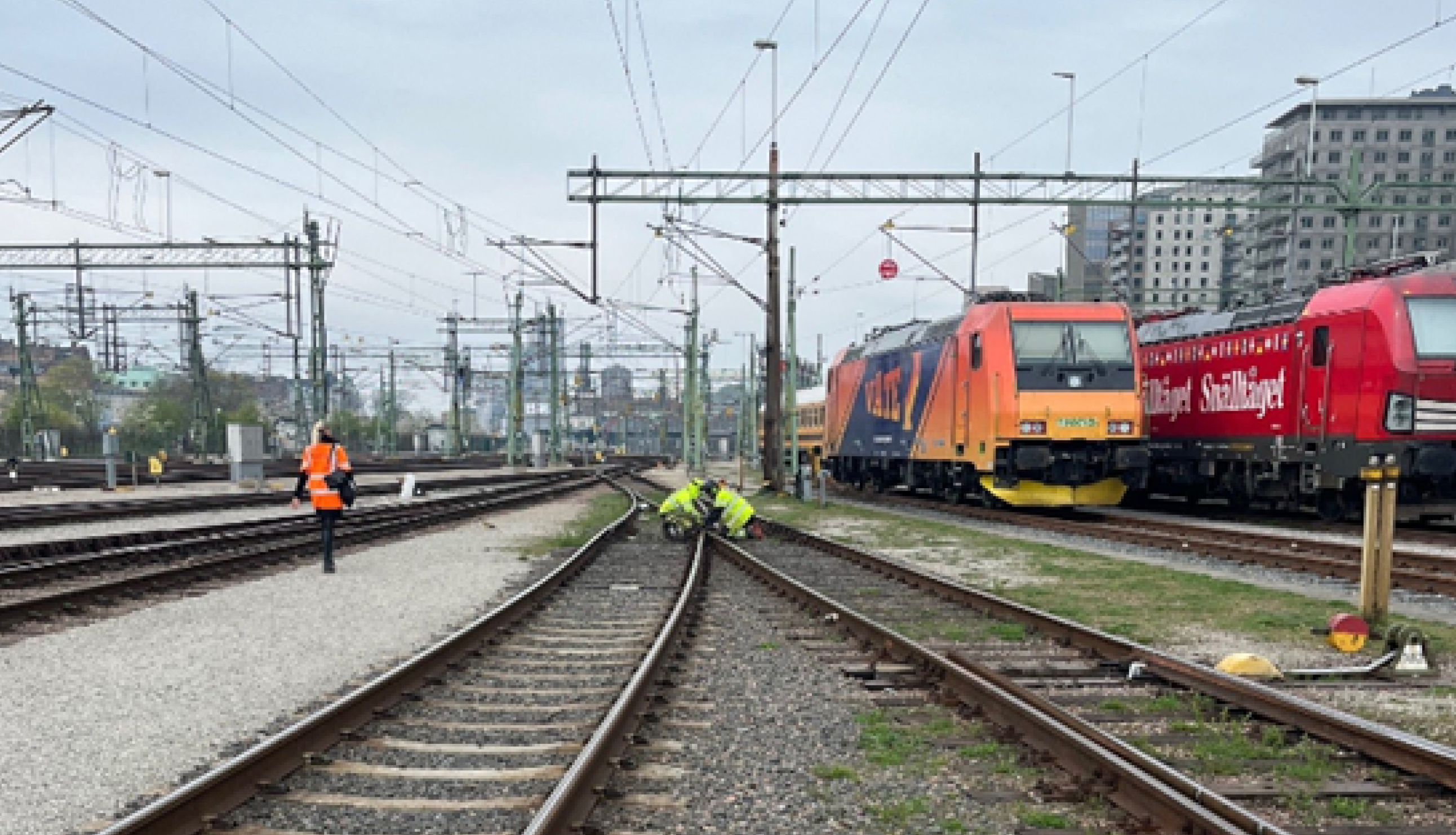 Arbetare i varselvästar granskar järnvägsspår vid en bangård, med två tåg intill och synliga ledningar ovanförför.