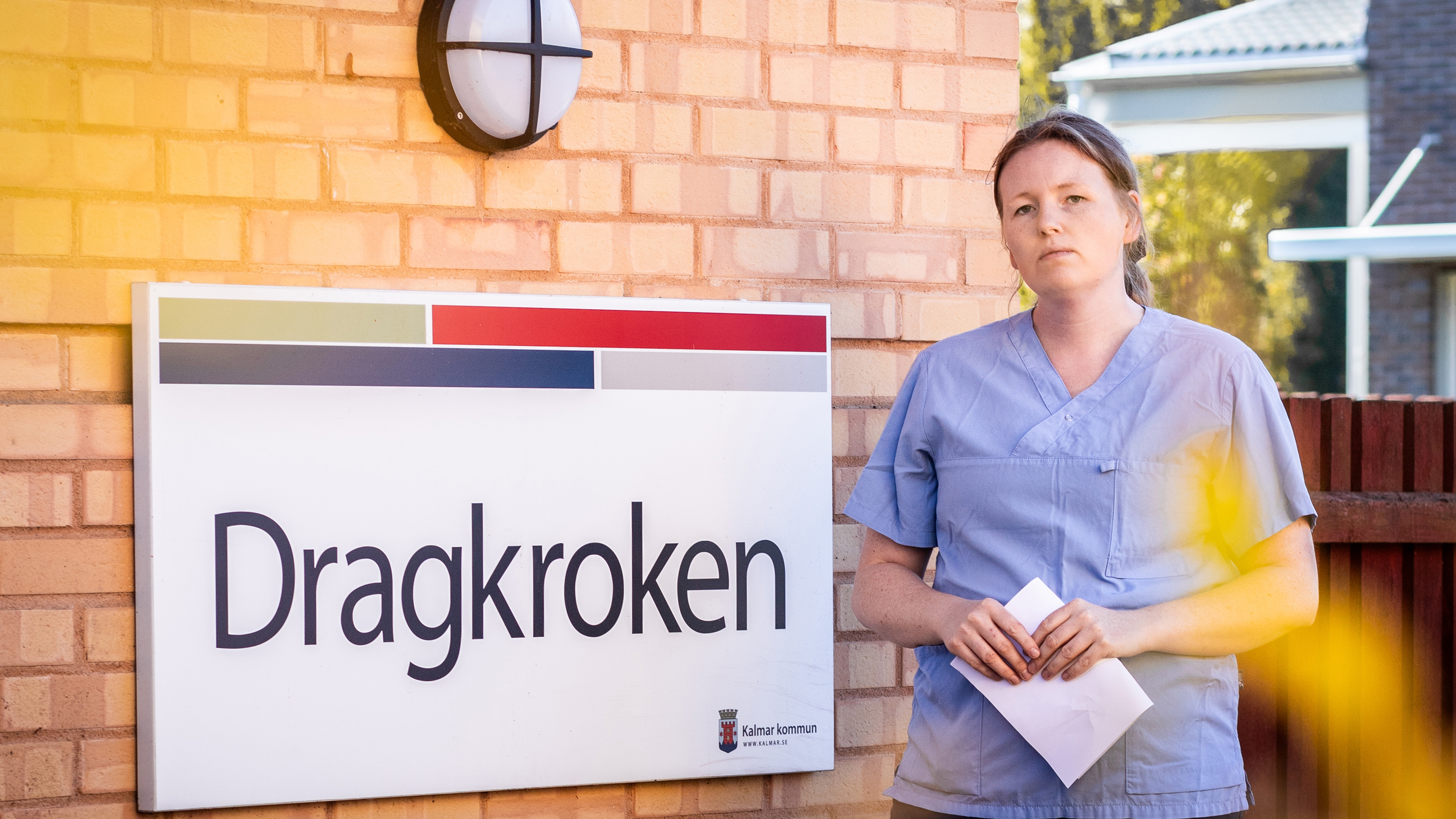 En kvinna i sjukhuskläder står bredvid en tegelvägg med en skylt där det står "Dragkroken". Hon håller papper och tittar mot kameran.