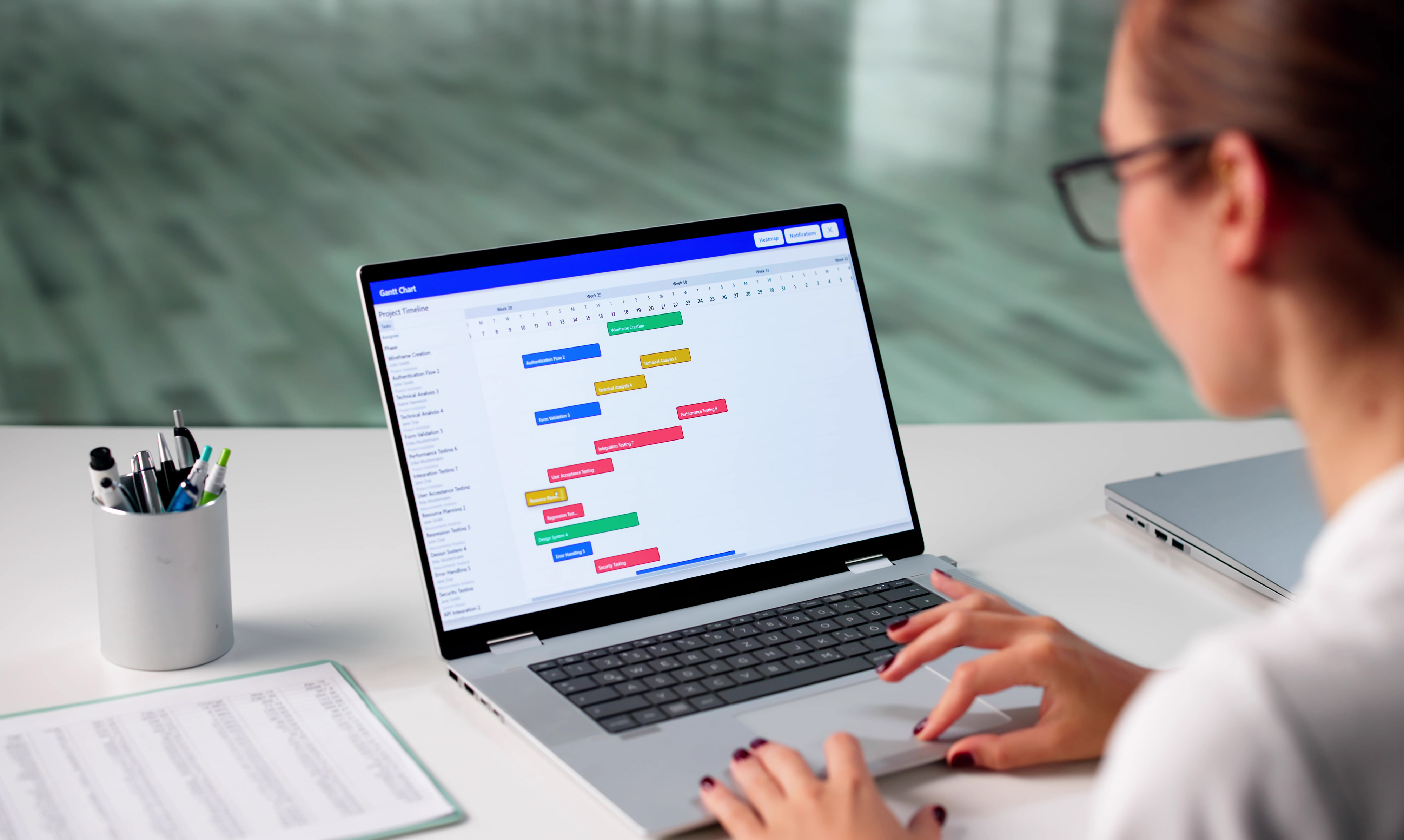 En person arbetar vid ett skrivbord med kontorsmaterial och har en bärbar dator som visar ett schema- eller projektplaneringsdiagram.