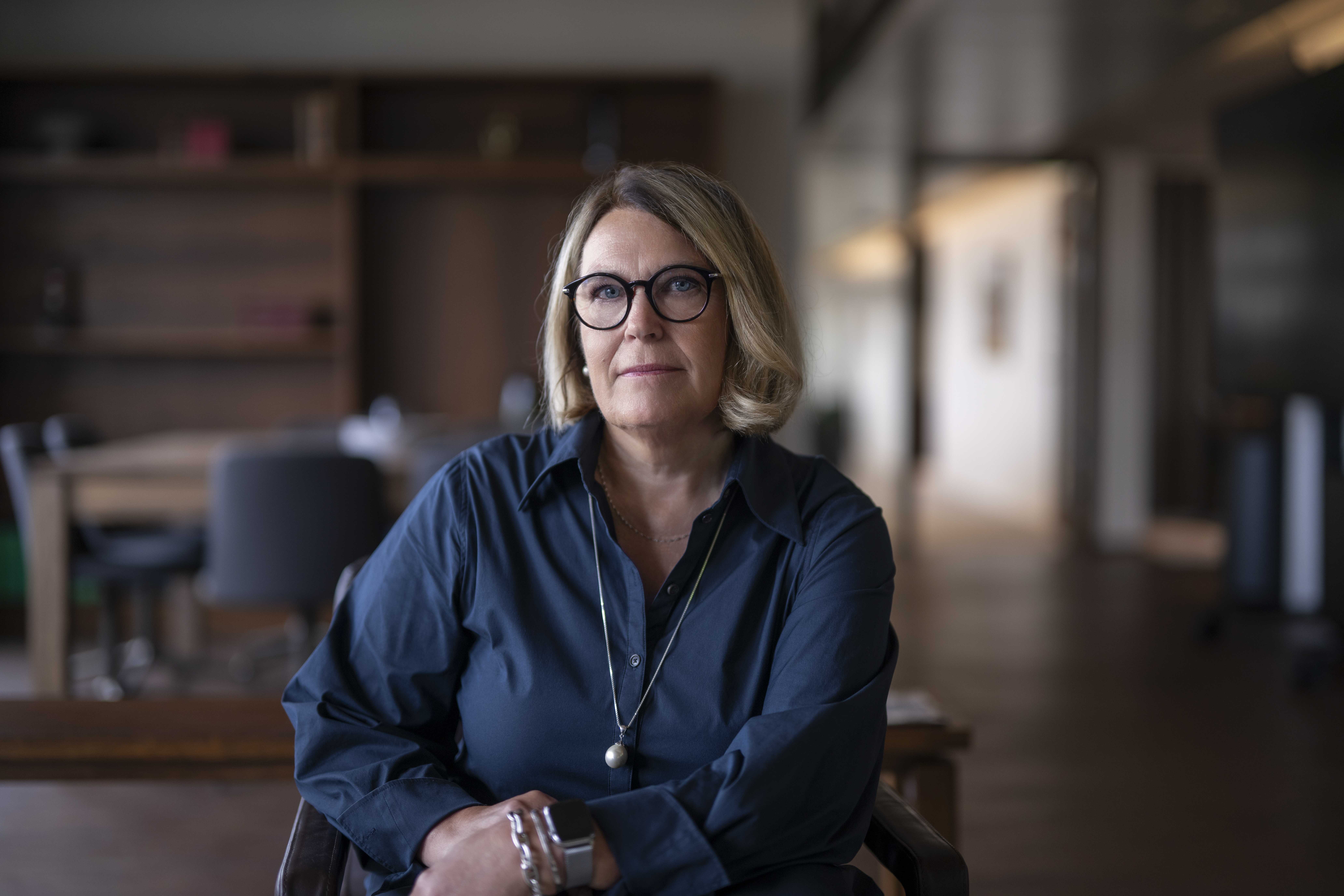 En medelålders kvinna med blont hår och glasögon sitter inomhus i en mörkblå tröja. Hyllor och bord syns i bakgrunden.