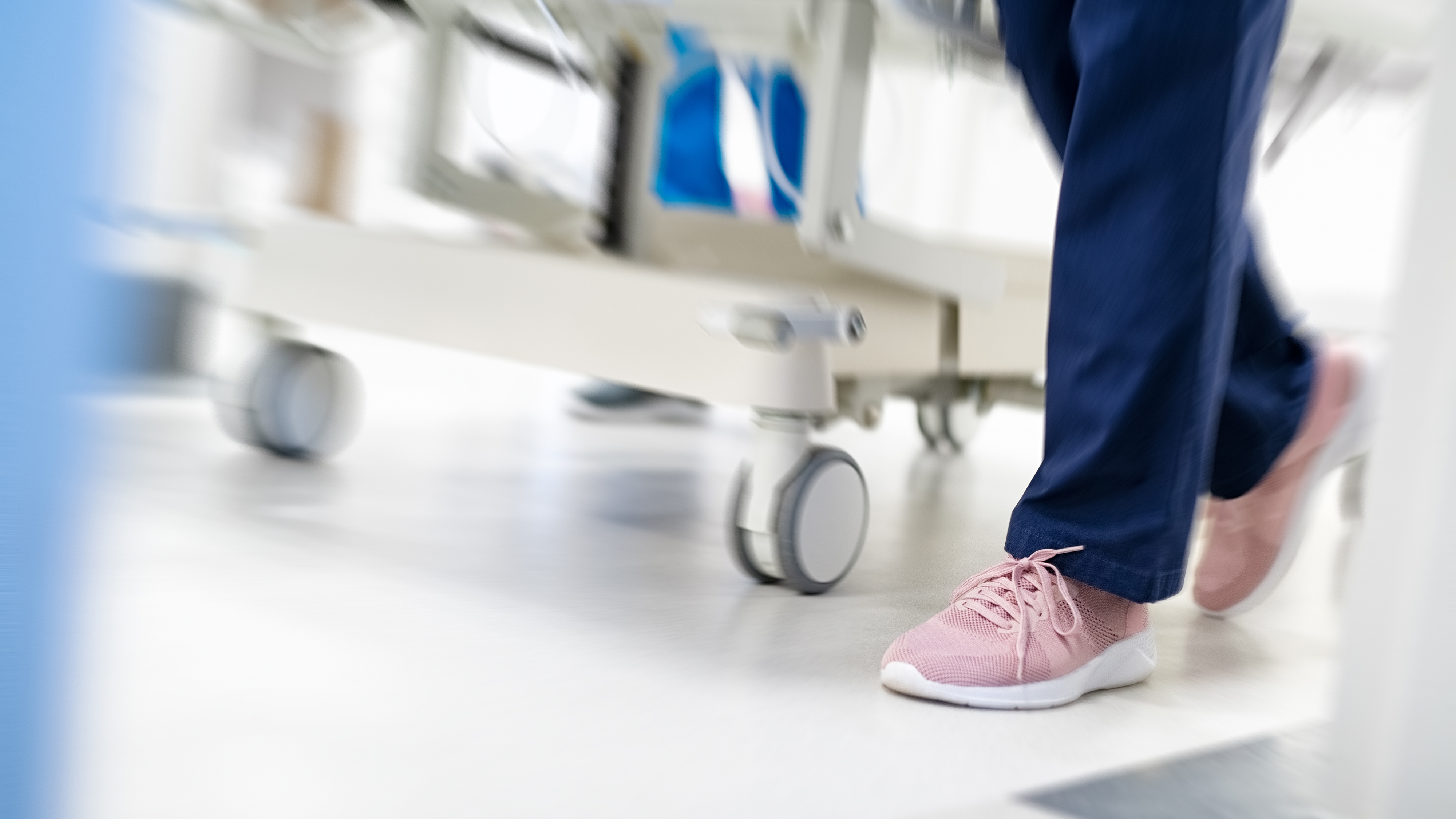 Närbild av en person med rosa sneakers och mörkblå byxor som går bredvid en sjukhussäng med hjul i en klinisk miljö.
