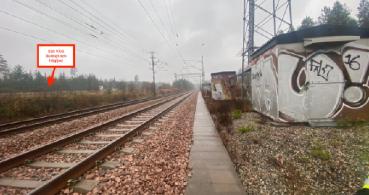 Två järnvägsspår löper parallellt bredvid en smal stig, med elstolpar, molnig himmel och graffiti på en byggnad. En röd skylt syns till vänster.