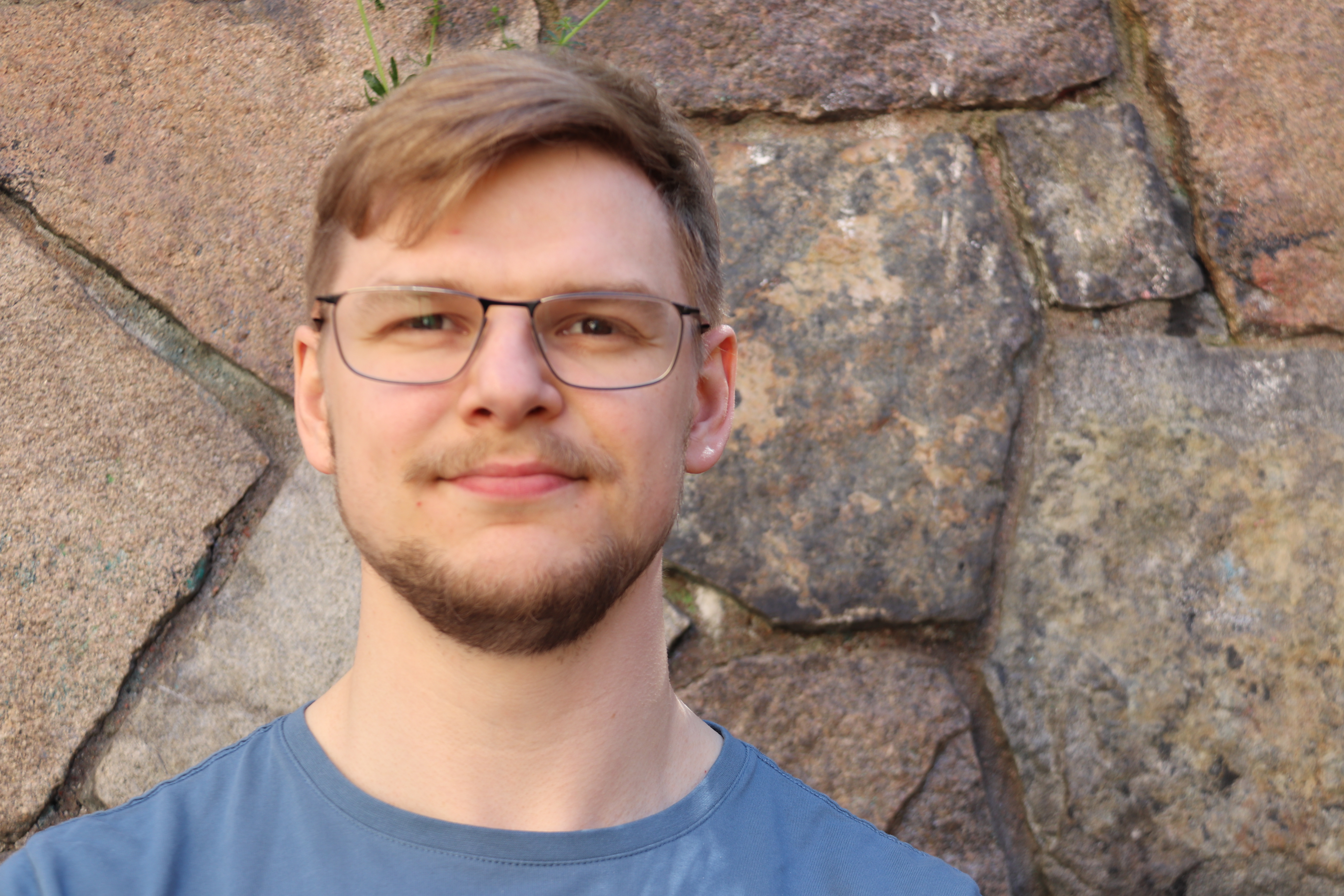 En man med kort blont hår, glasögon och ljus skäggväxt står framför en stenvägg och har på sig en ljusblå skjorta.