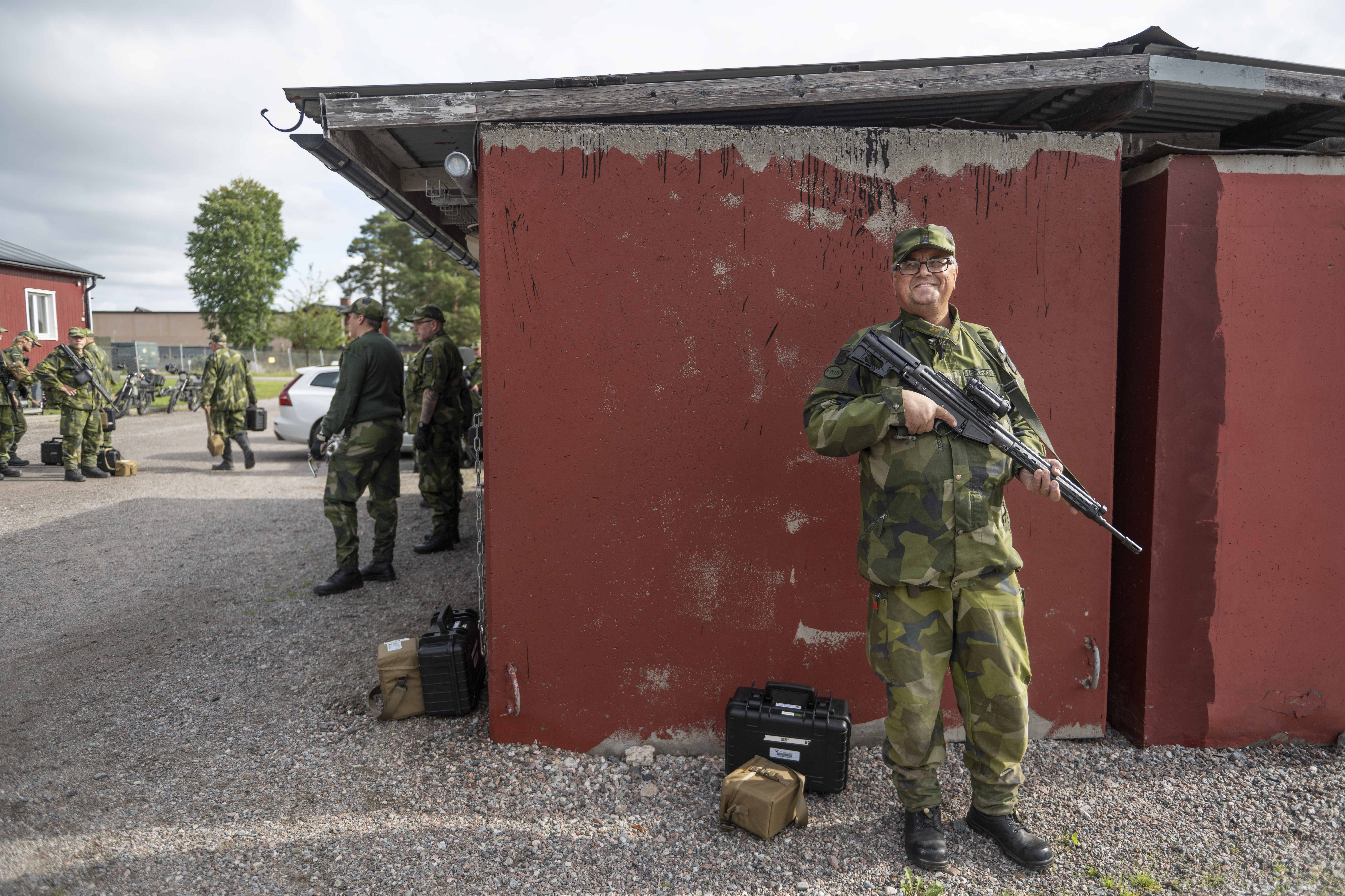 En soldat i kamouflageuniform står vakt med ett gevär vid en röd byggnad; flera andra soldater syns i bakgrunden.