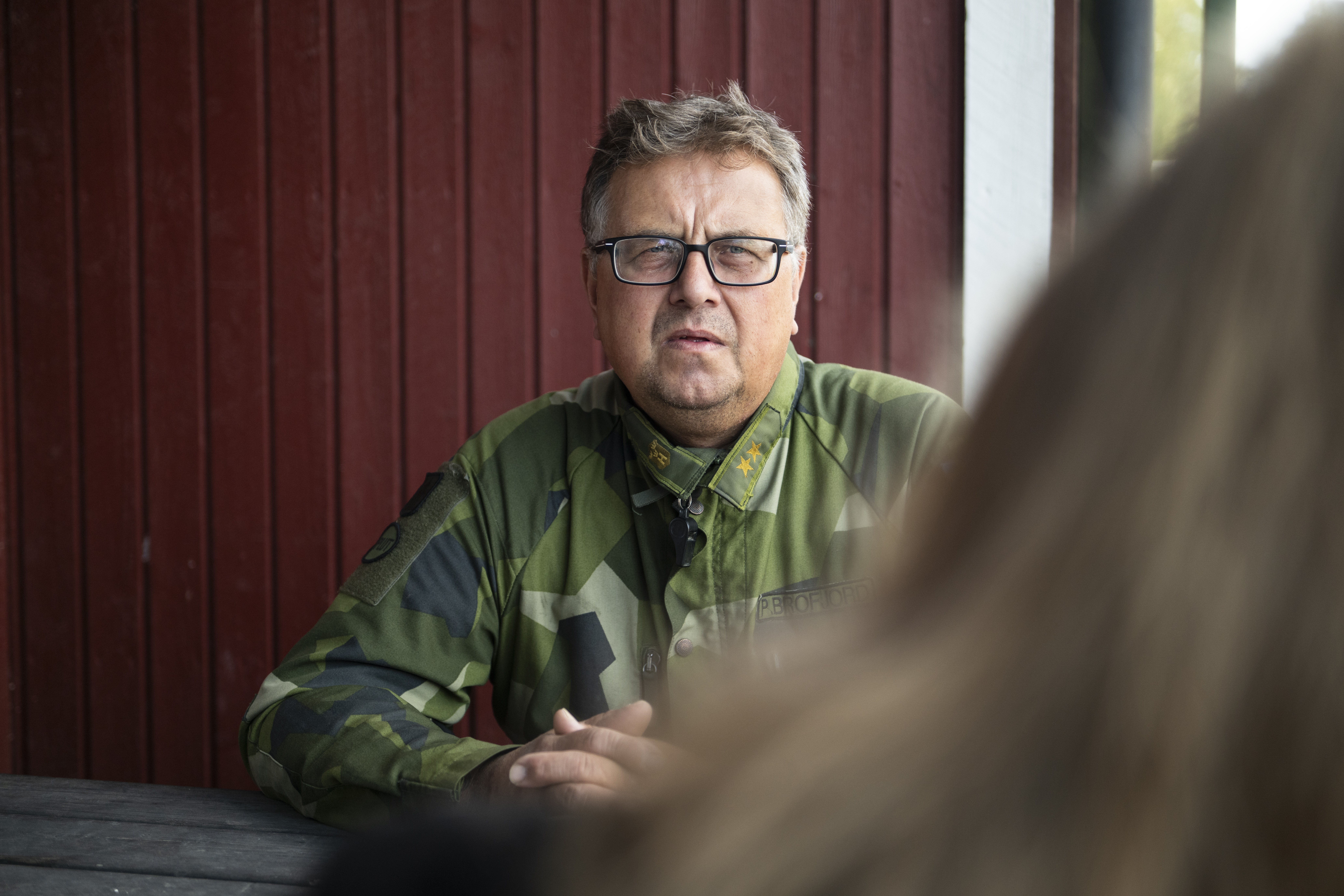 En man i militäruniform sitter vid ett bord framför en röd trävägg och tittar framåt. En annan person syns delvis i förgrunden.