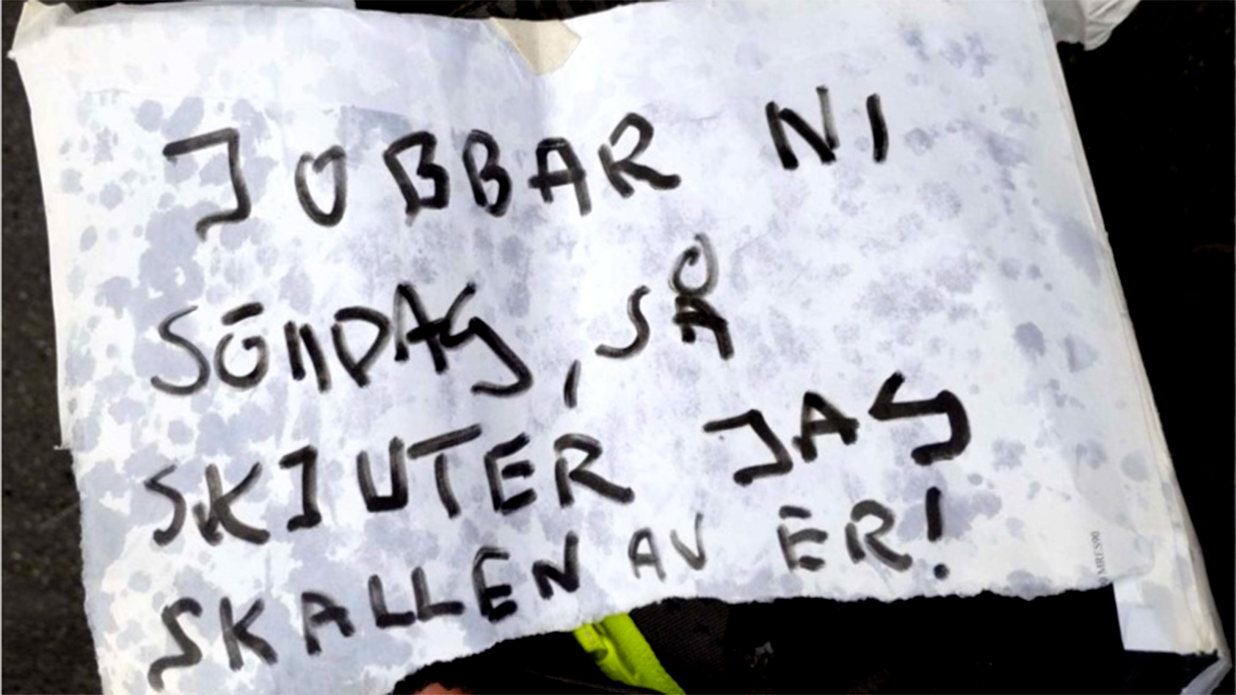En handskriven skylt på svenska lyder: "JOBBAR NI SÖNDAG, SÅ SKJUTER JAG SKALLEN AV ER!" på ett papper med veck.