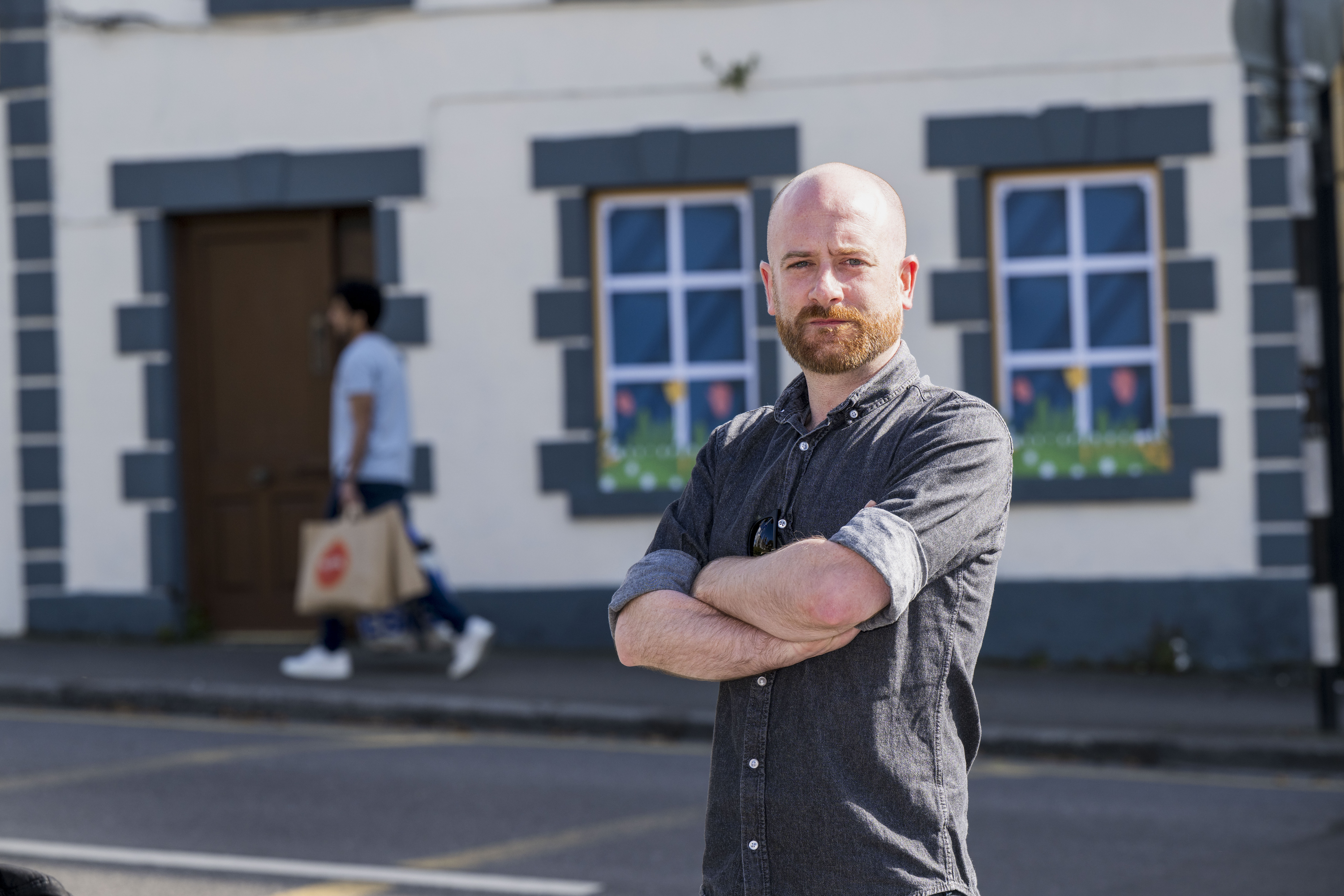 Eoghan Fox med skägg och korsade armar står på en gata framför en byggnad med blå fönster; en annan person går förbi med en shoppingkasse.