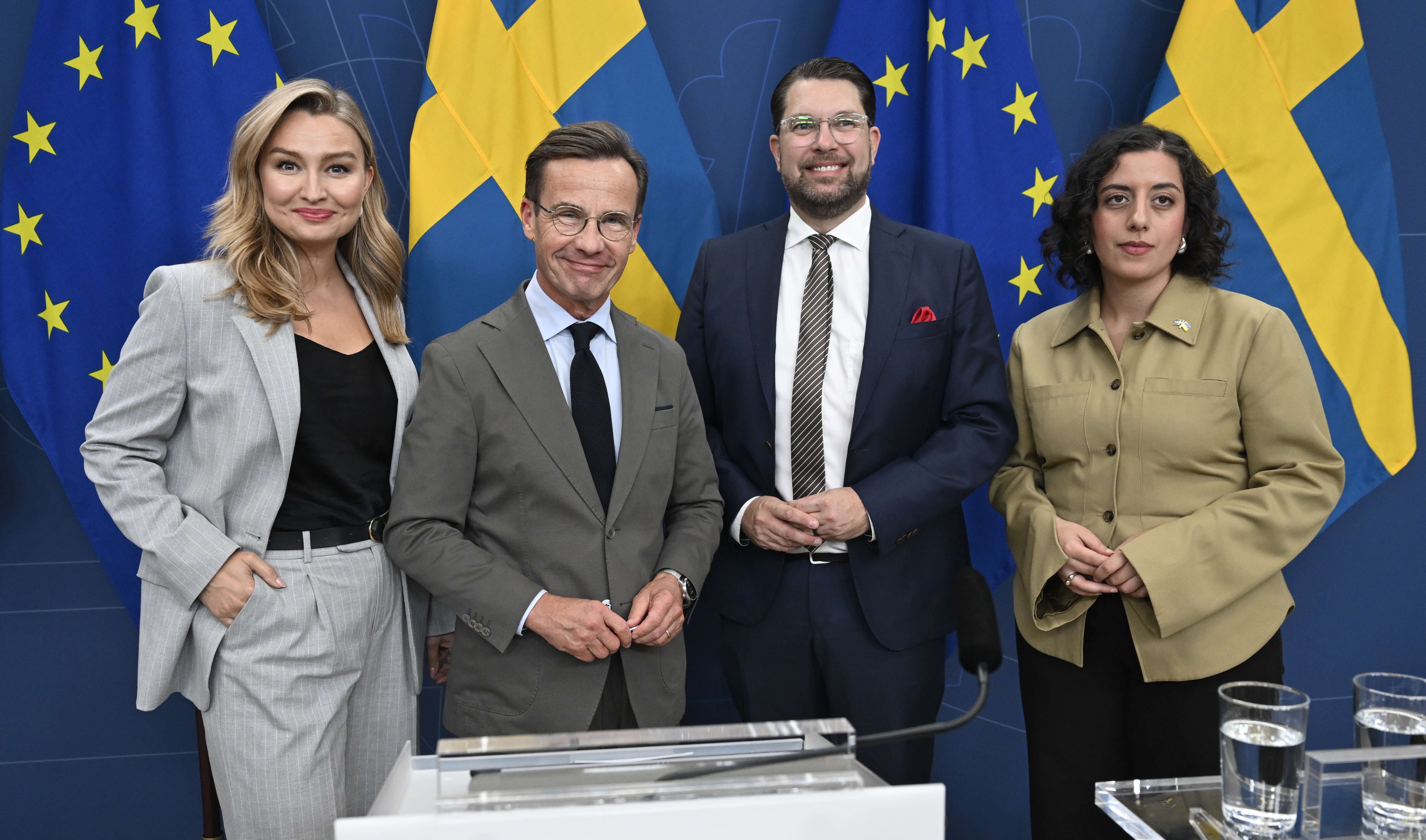 Fyra personer i affärskläder står framför EU:s och Sveriges flaggor och poserar för en gruppbild vid en pressträff.