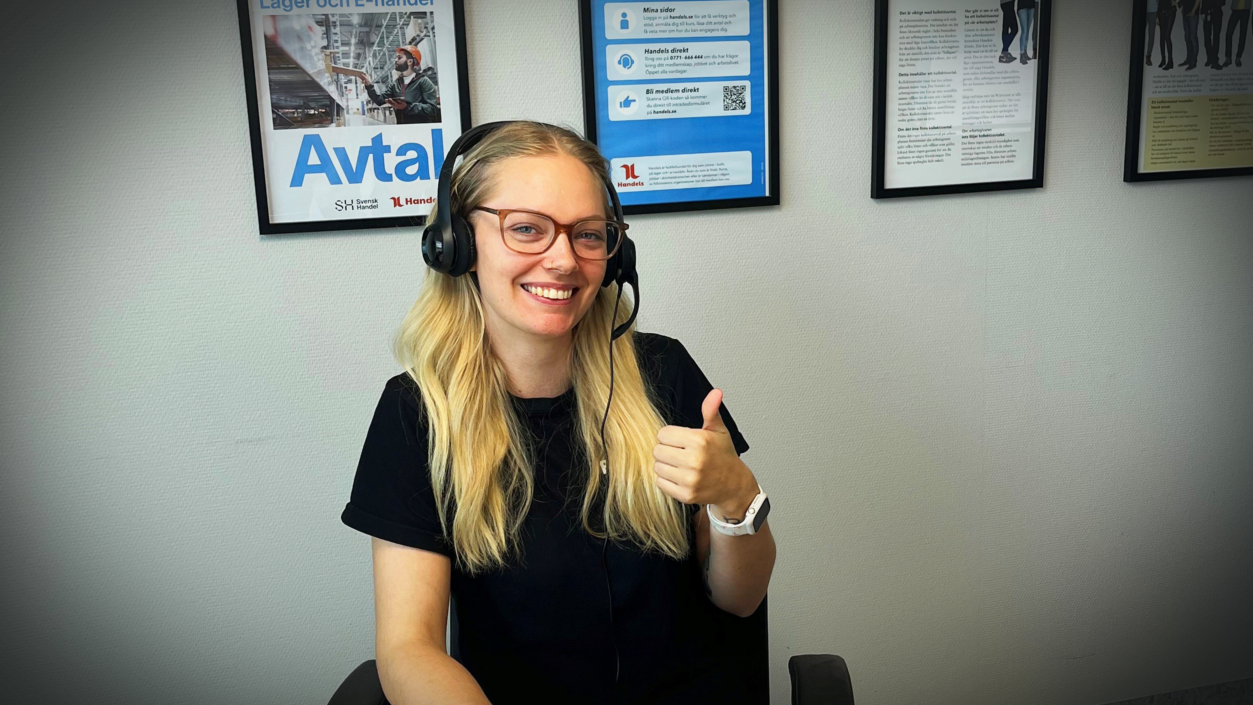 En kvinna med långt blont hår, glasögon och headset sitter vid ett skrivbord och gör en tumme upp. Informationsaffischer är på väggen bakom henne.