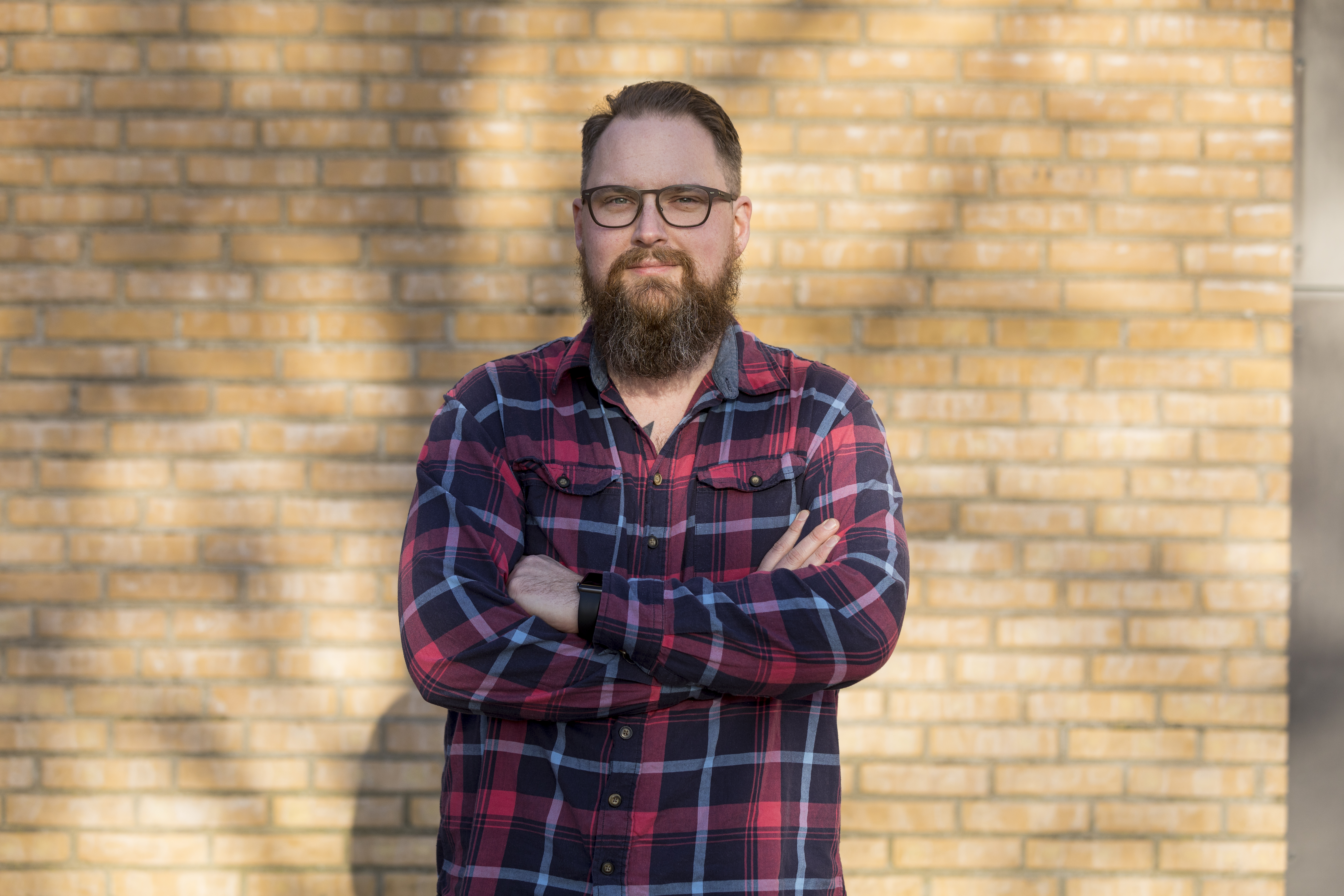 En man med glasögon och skägg står med armarna i kors framför en tegelvägg, iförd en röd och blå rutig skjorta.