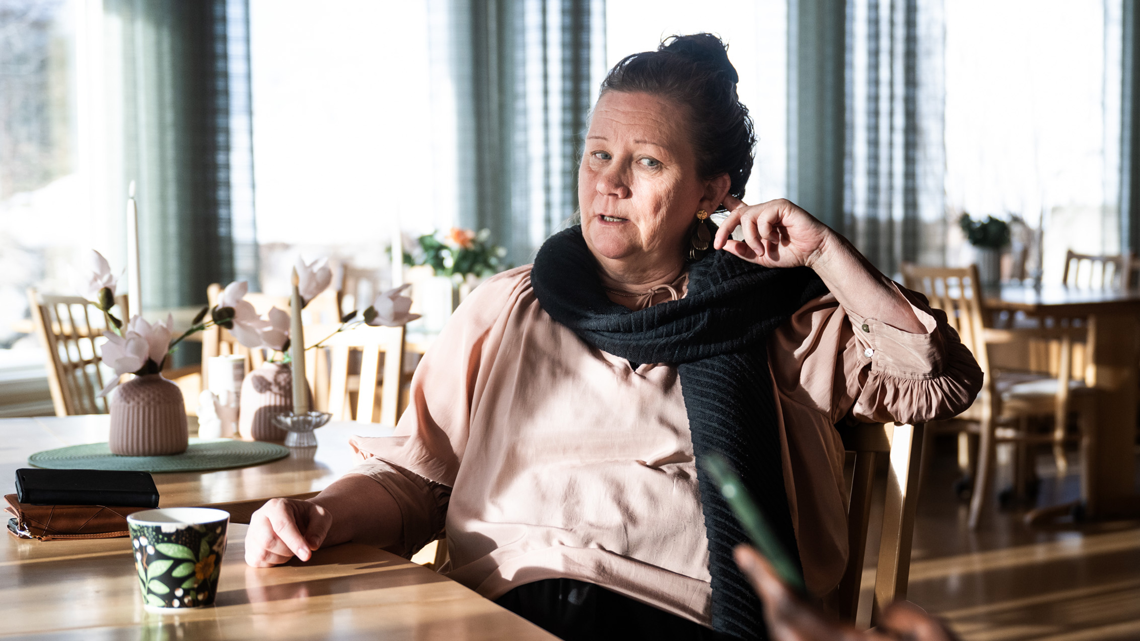 En kvinna sitter vid ett bord av trä i ett ljust rum, har en ljus blus och mörk sjal. Handen är vid örat. Blommor och koppar står på bordet.