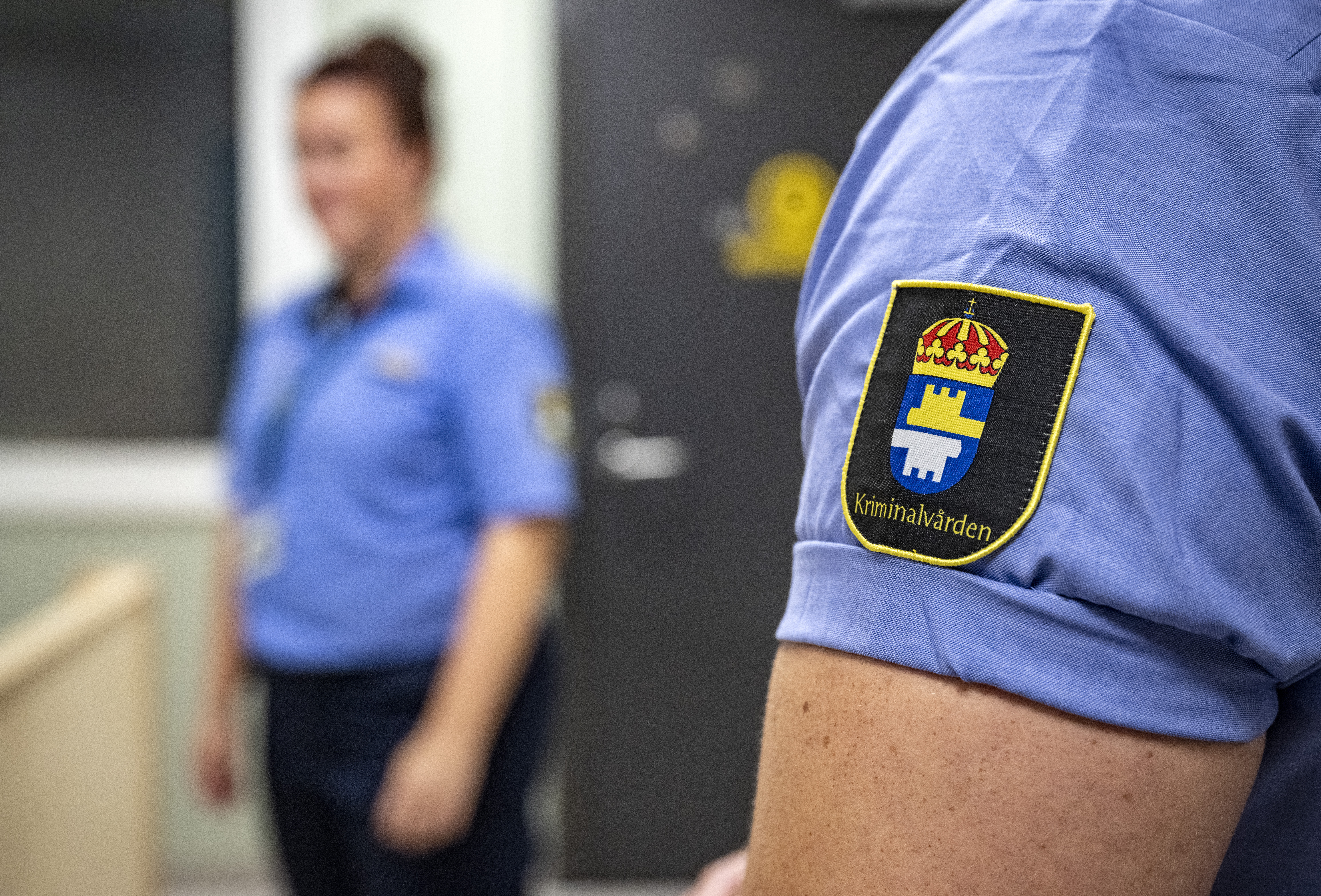 En person i blå uniform med "Kriminalvården"-märke. En annan person i liknande uniform står i bakgrunden inomhus.