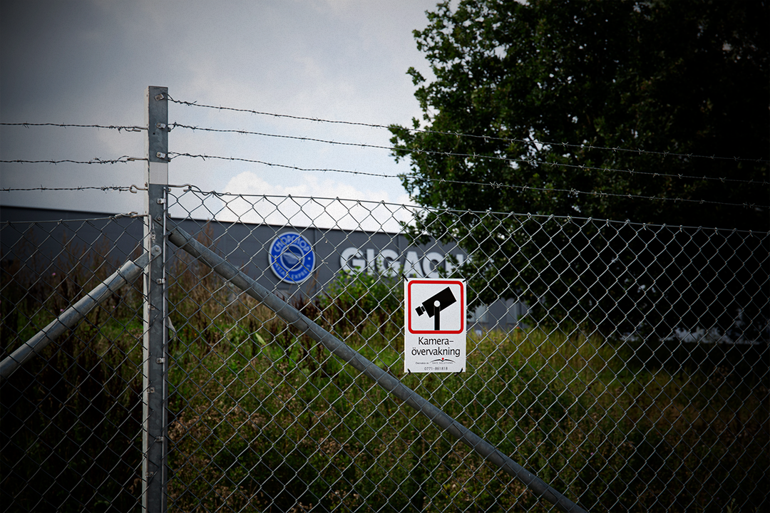 Ett stängsel av metallnät med taggtråd och en skylt om kameraövervakning framför en industribyggnad och träd.