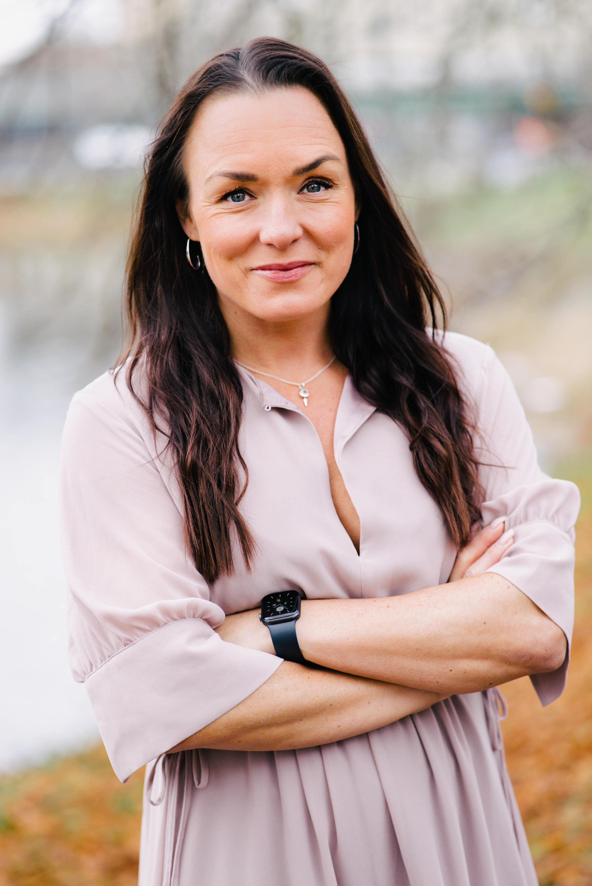 Linda Larsson med långt brunt hår, ljusrosa klänning och svart smartklocka står utomhus med armarna i kors och neutral min.