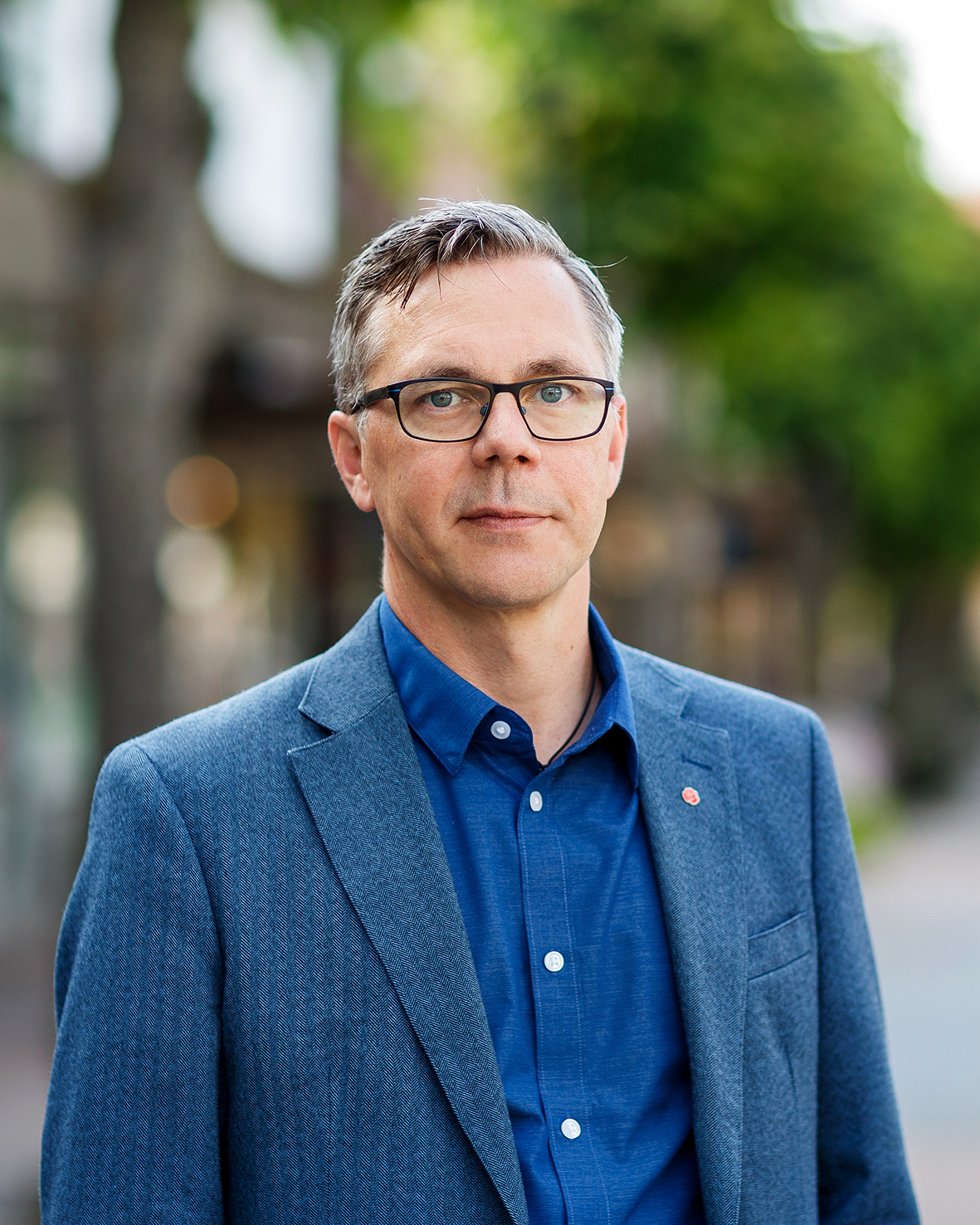 Johan Söderberg med glasögon och blå kavaj över en blå skjorta står utomhus med träd och byggnader i bakgrunden.