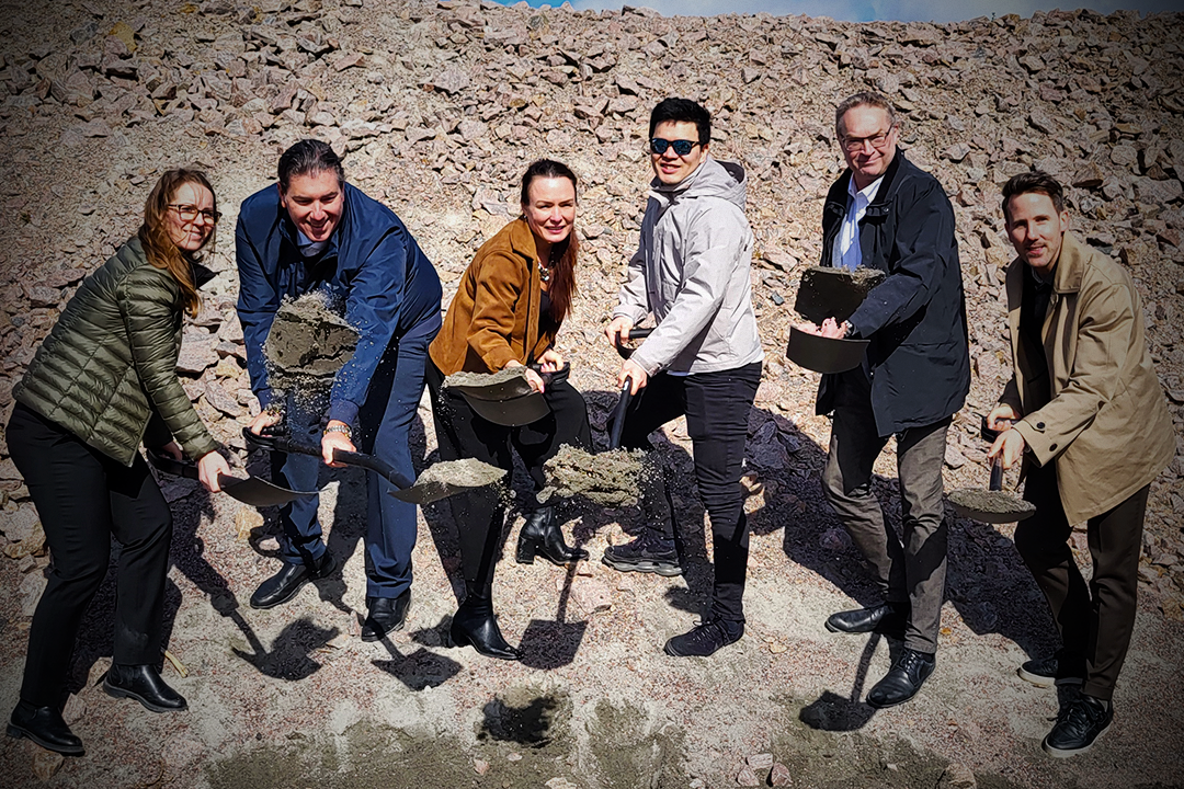 Sex personer står på rad utomhus, alla håller en spade med jord och verkar delta i en första spadtagsceremoni.