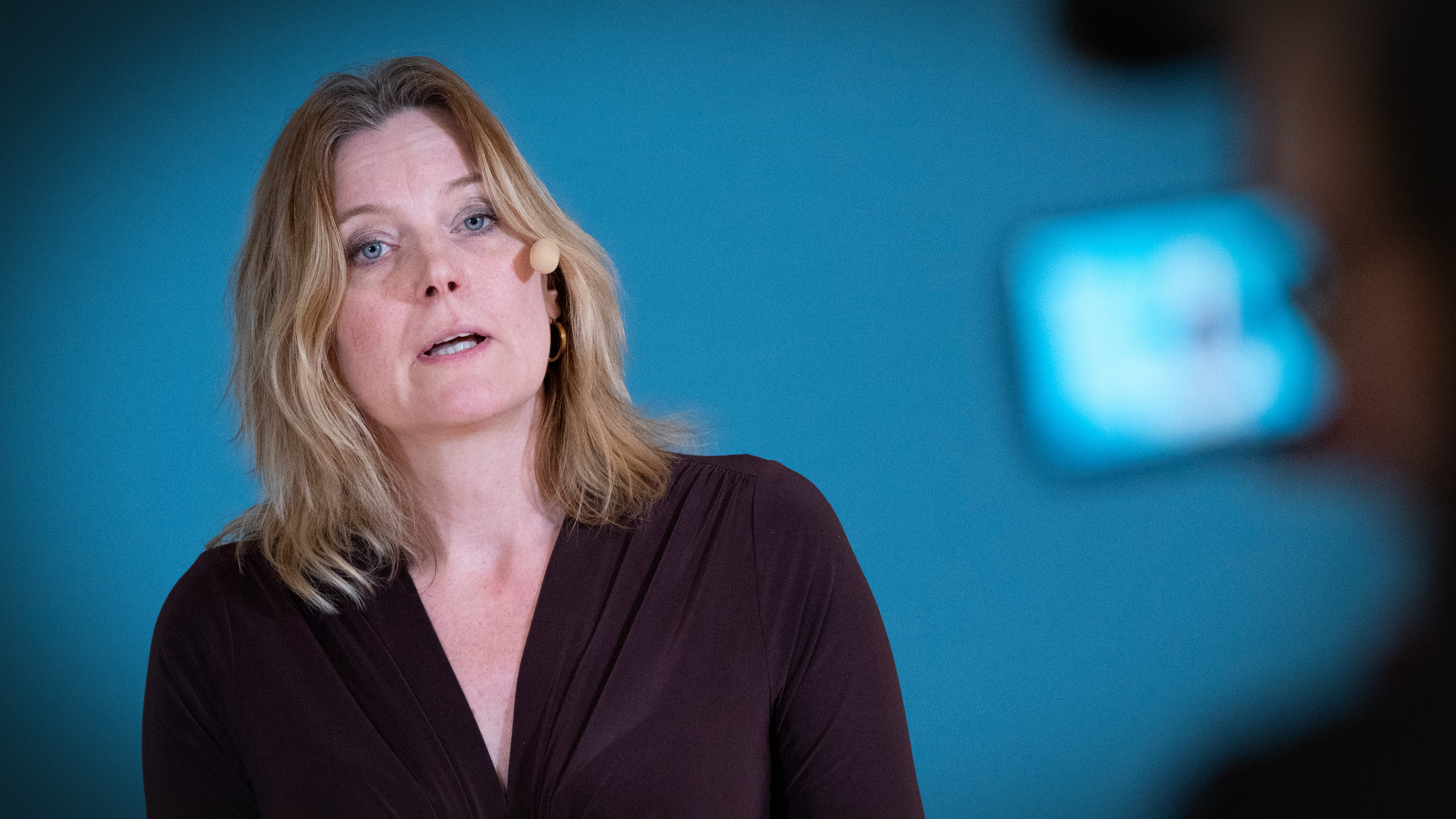 Ulrika Geeraedts med blont hår och headsetmikrofon talar framför en blå bakgrund, medan en kamera filmar henne i förgrunden.