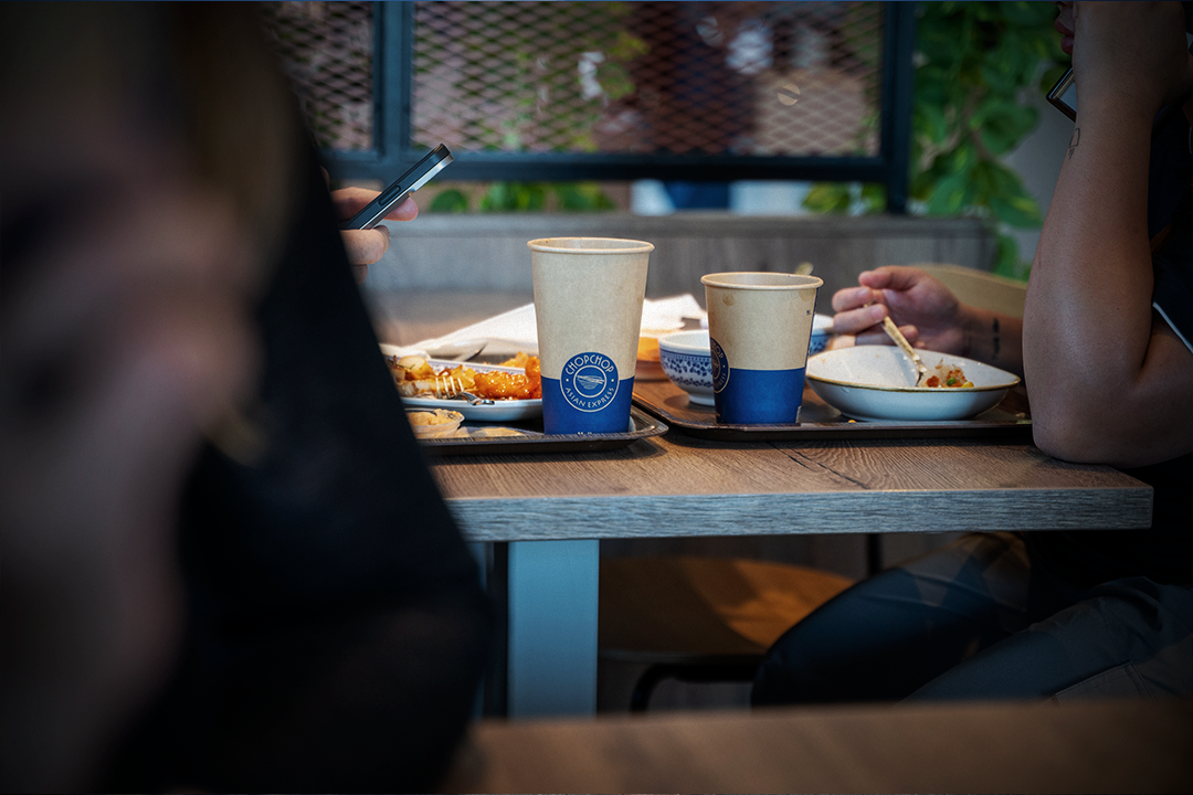 Två personer sitter vid ett bord med engångsmuggar och skålar med matte; en person använder en smartphone.