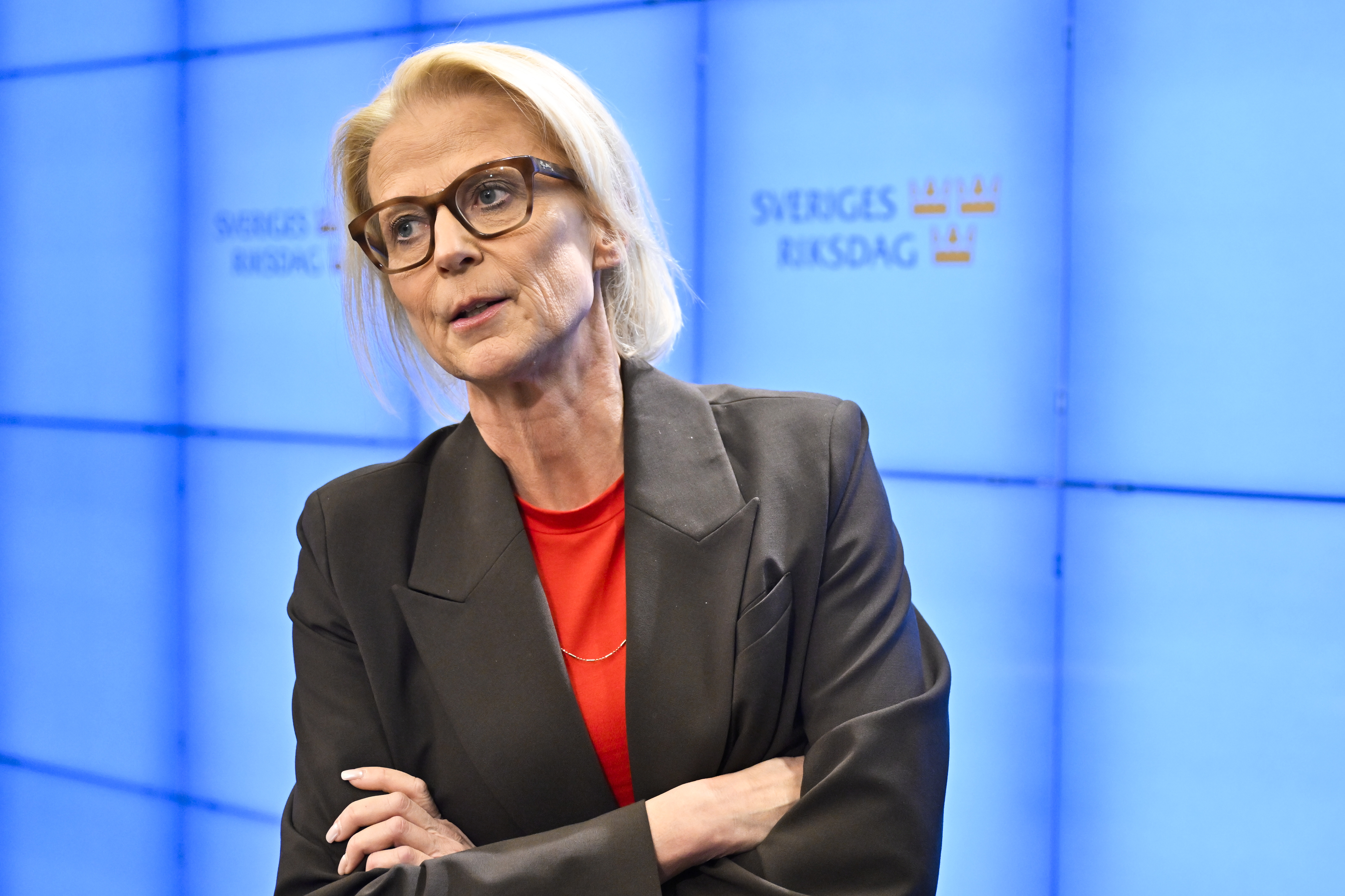 En kvinna med kavaj och röd skjorta står med armarna i kors framför en blå bakgrund med texten "Sveriges Riksdag" och logotyper.