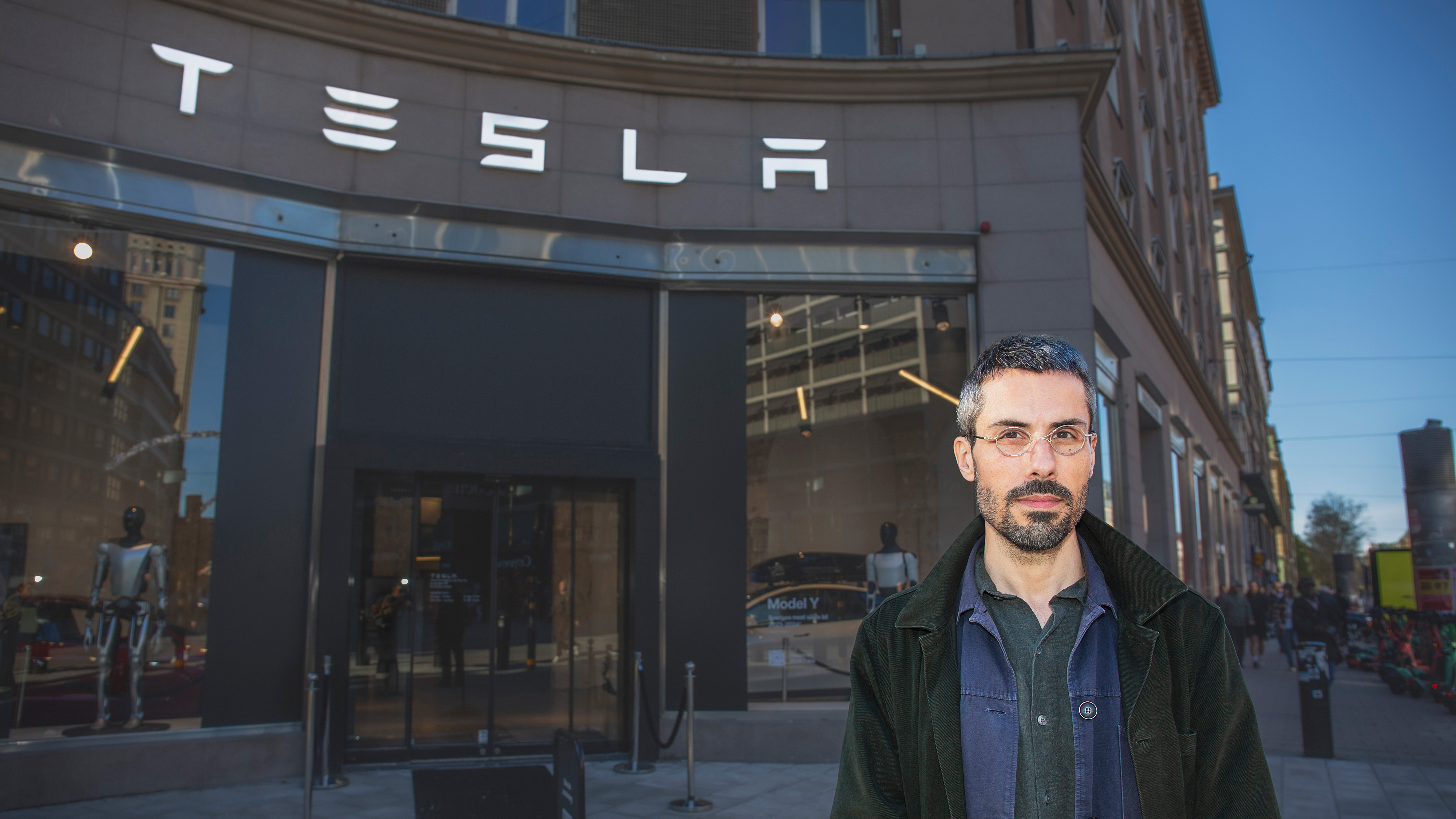 En man med glasögon och skägg står framför en Tesla-butik med stora fönster och Teslas logotyp på byggnaden.