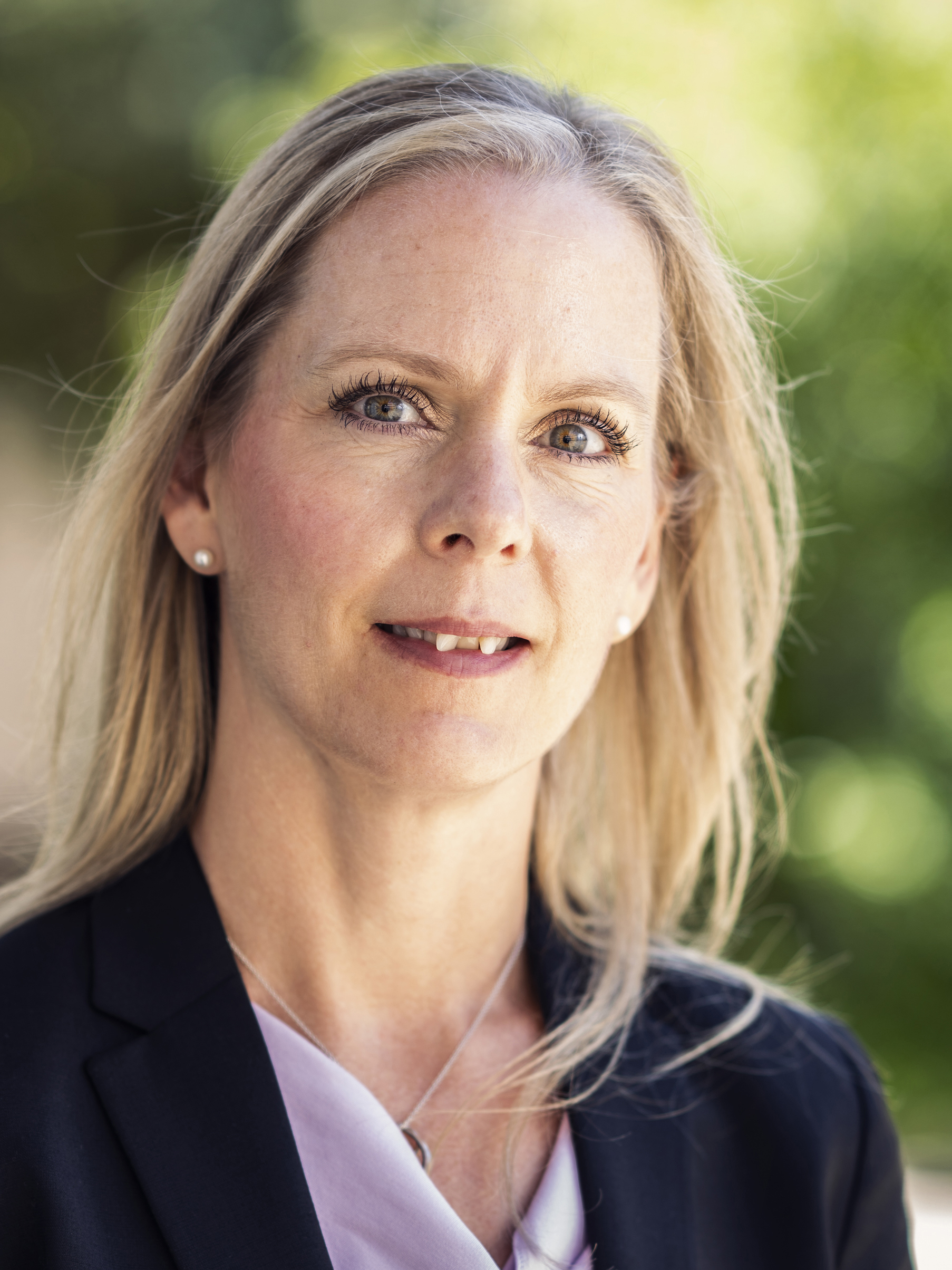 Ankie Hartzén med svart kavaj och ljus blus står utomhus och tittar in i kameran, med grön bakgrund i oskärpa.