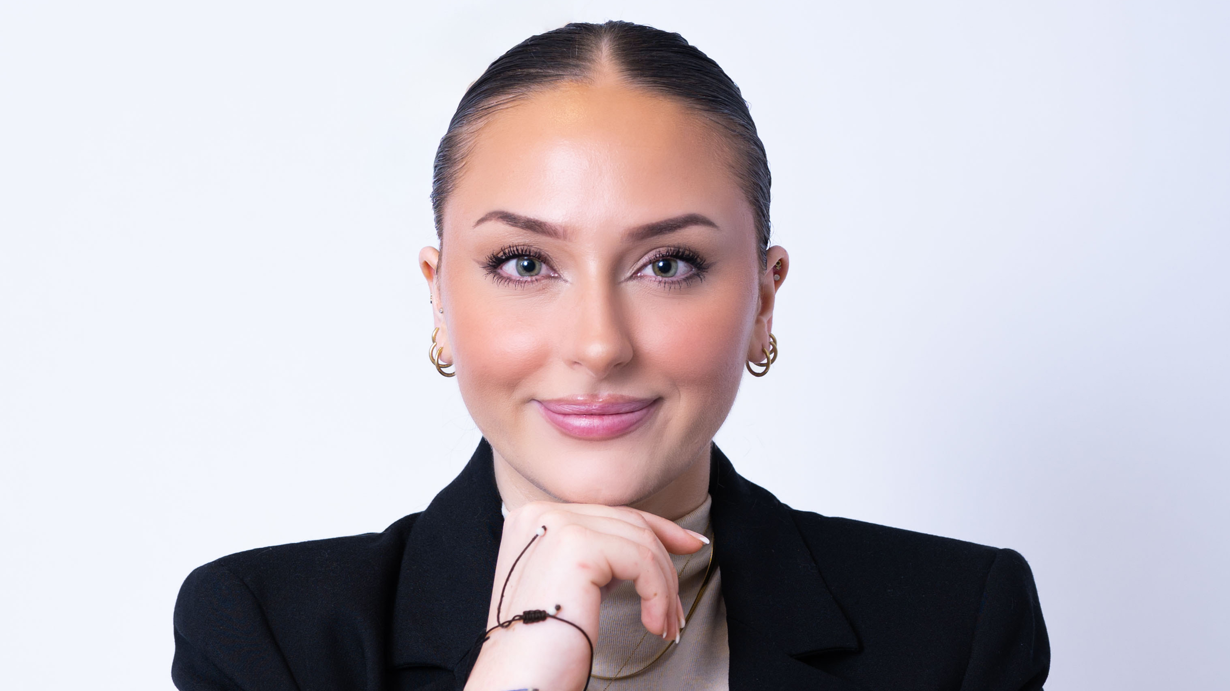 En kvinna i svart kavaj vilar hakan på handen och ler svagt mot en ljus enfärgad bakgrund.