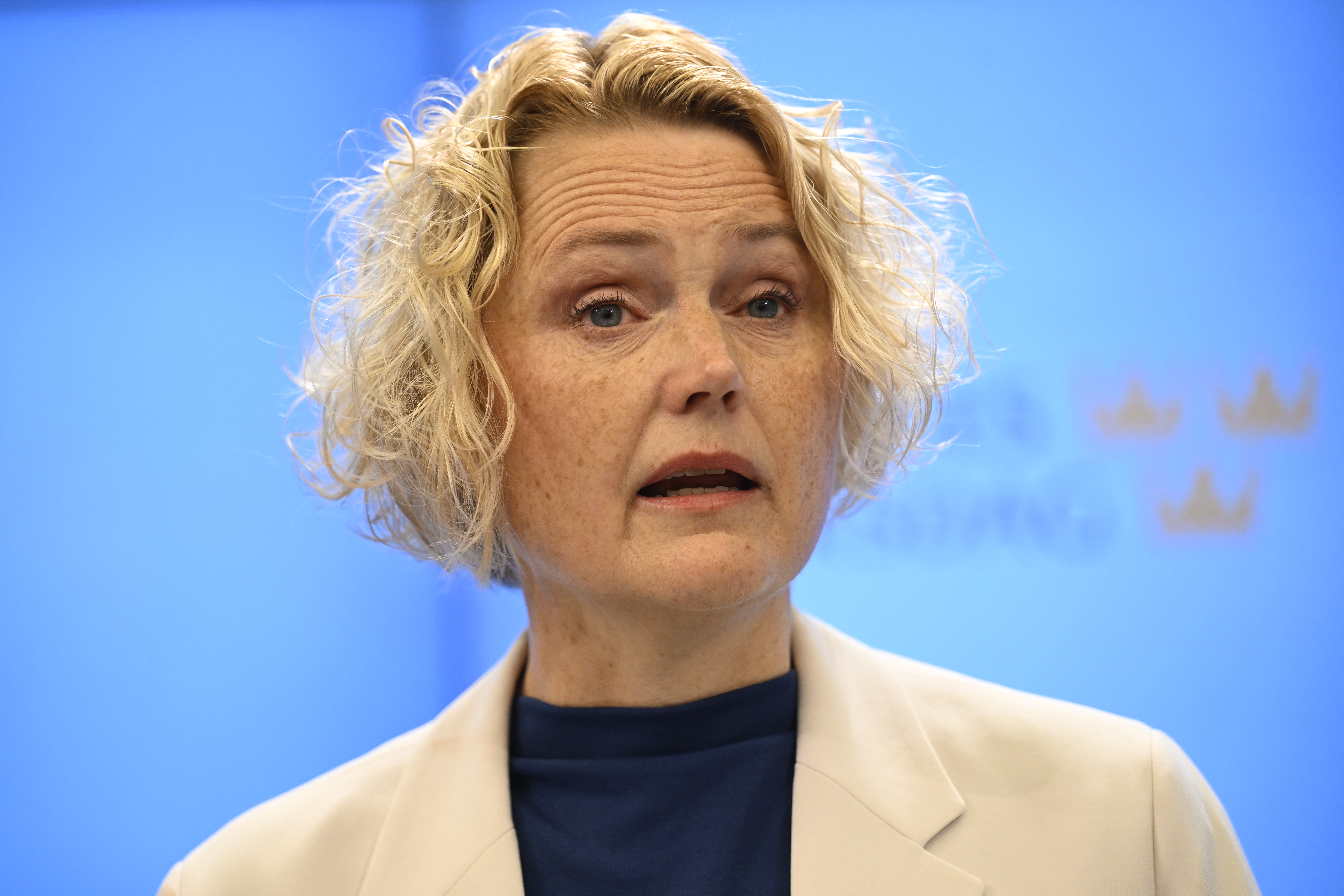 Anna-Karin Hatt med kort lockigt blont hår, ljus kavaj och mörk topp talar framför en blå bakgrund med svaga gula kronor.