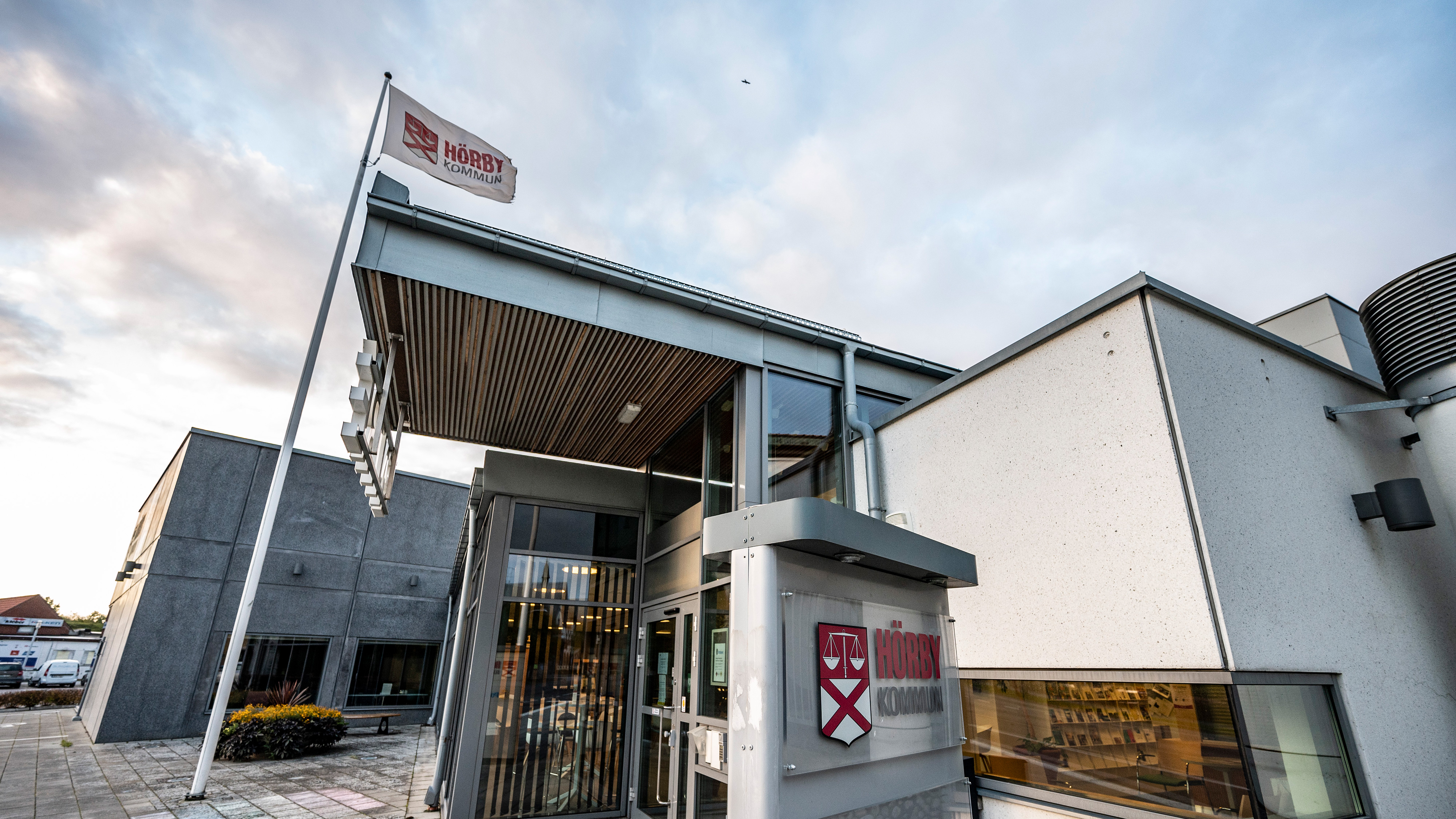 Byggnad med stora fönster, en flagga och en skylt med texten "Hörby kommun" ovanför ingången under molnig himmel.