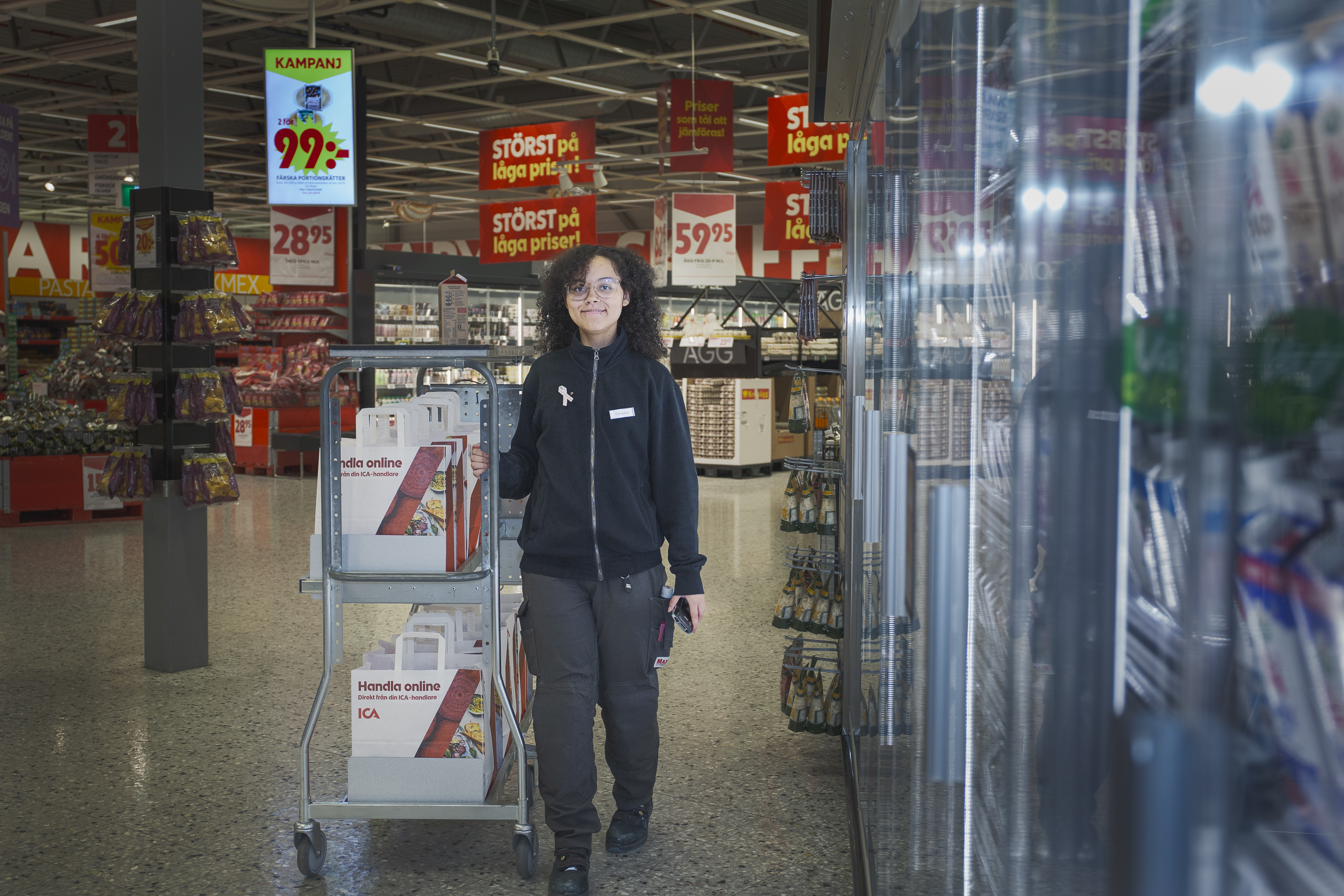 En person skjuter en vagn med kartonger genom en mataffärsgång med reklamskyltar och kyldiskar.