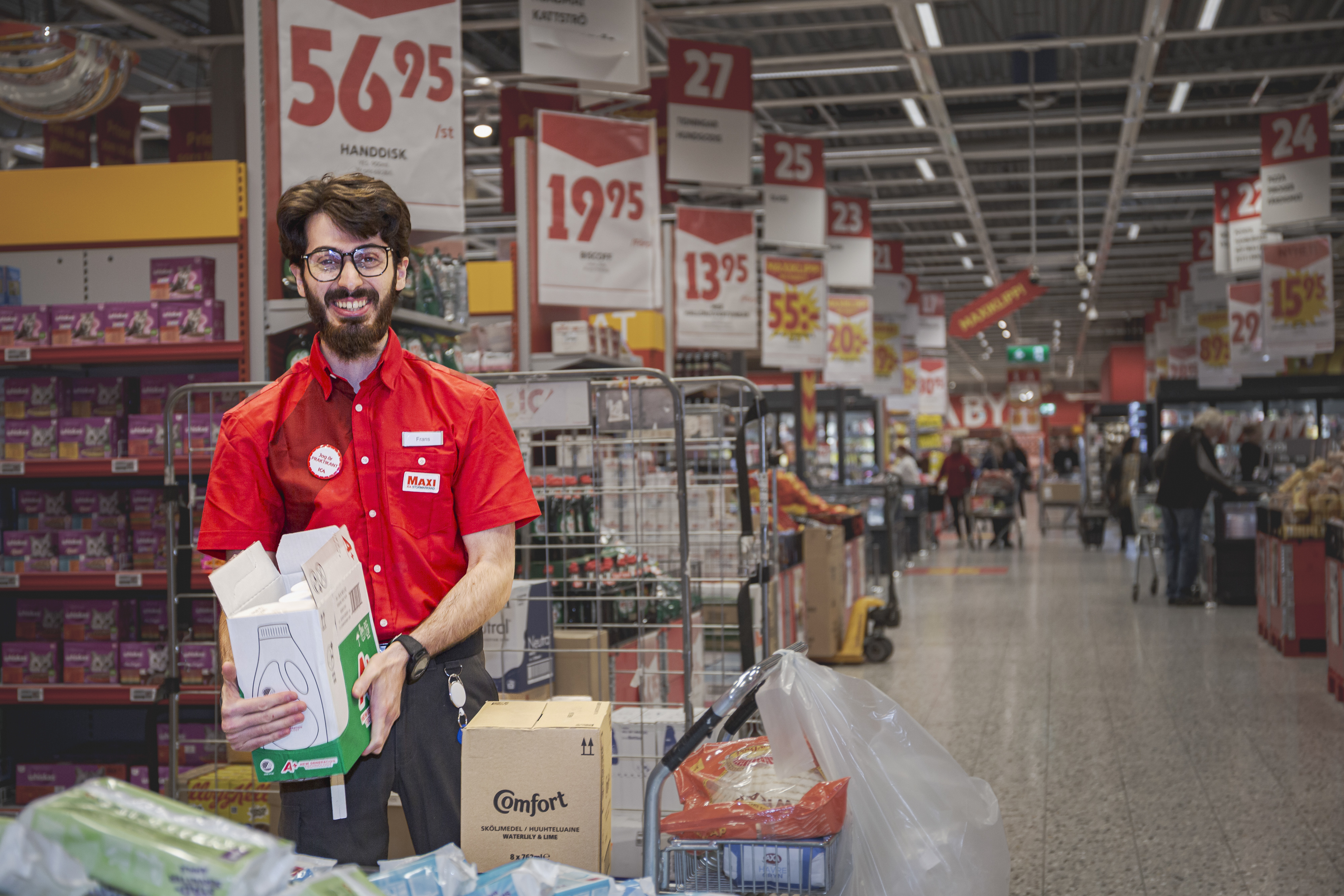 En butiksanställd i röd tröja står vid en vagn med lådor och plastpåsar i en butiksgång med prisskyltar ovanförför.
