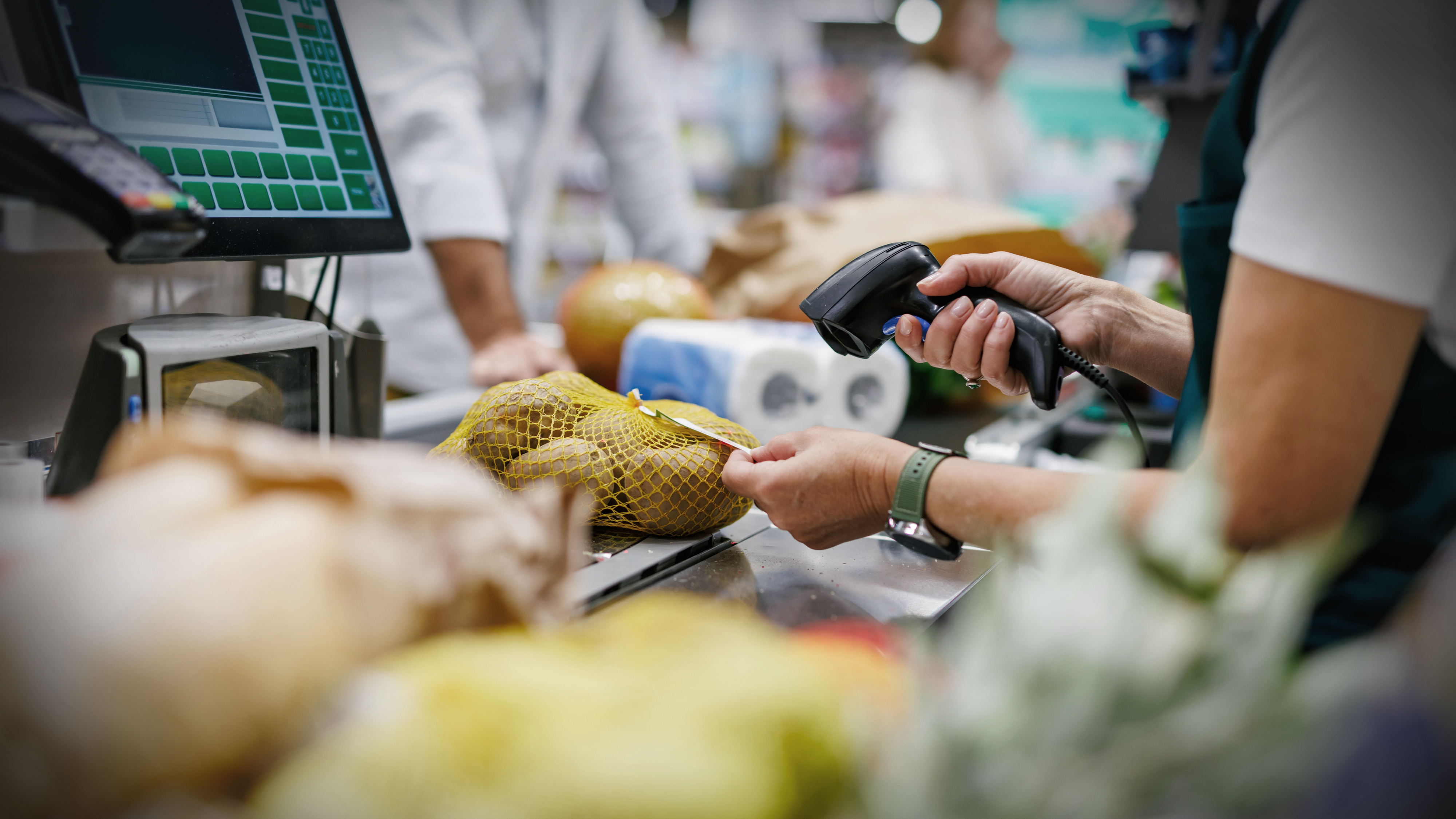 En kassör skannar en påse potatis i en butikskassa medan en datorskärm visar transaktionsuppgifter.