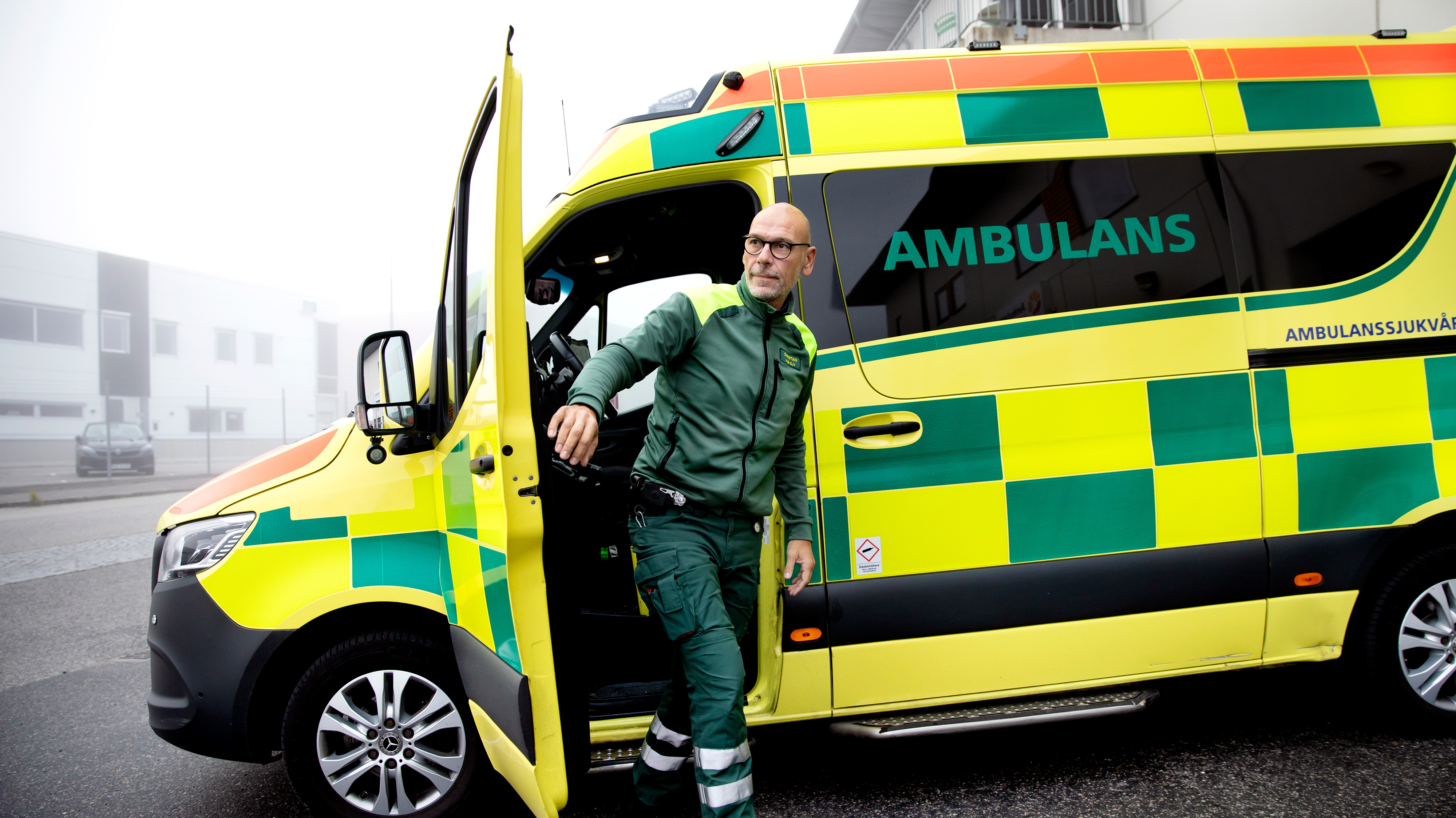 En ambulanssjukvårdare i uniform kliver ut ur en gul och grön ambulans parkerad utanför en byggnad en dimmig dag.