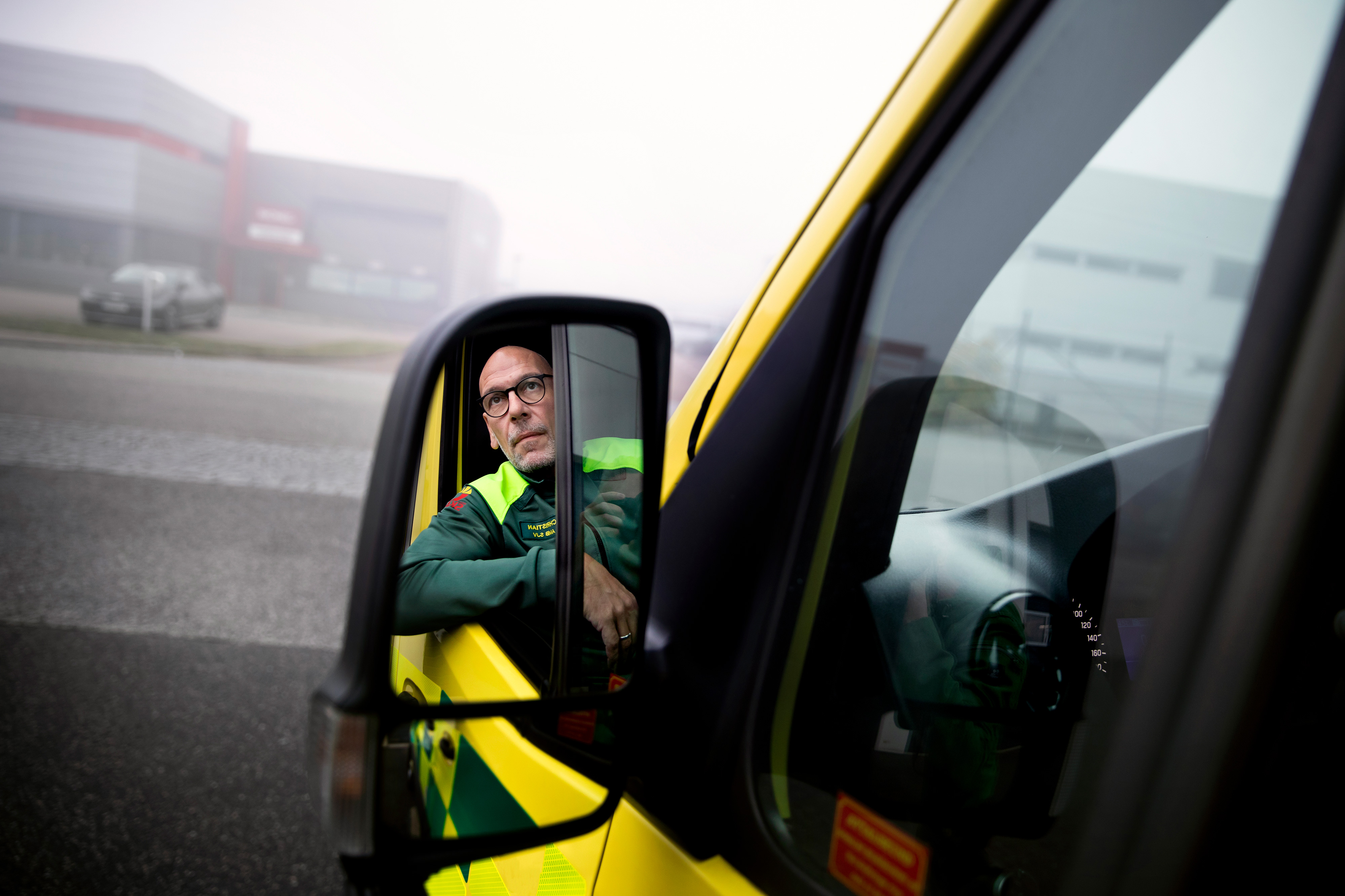 En man i arbetskläder syns i sidospegeln på ett gult utryckningsfordon som står parkerat på en dimmig gata.