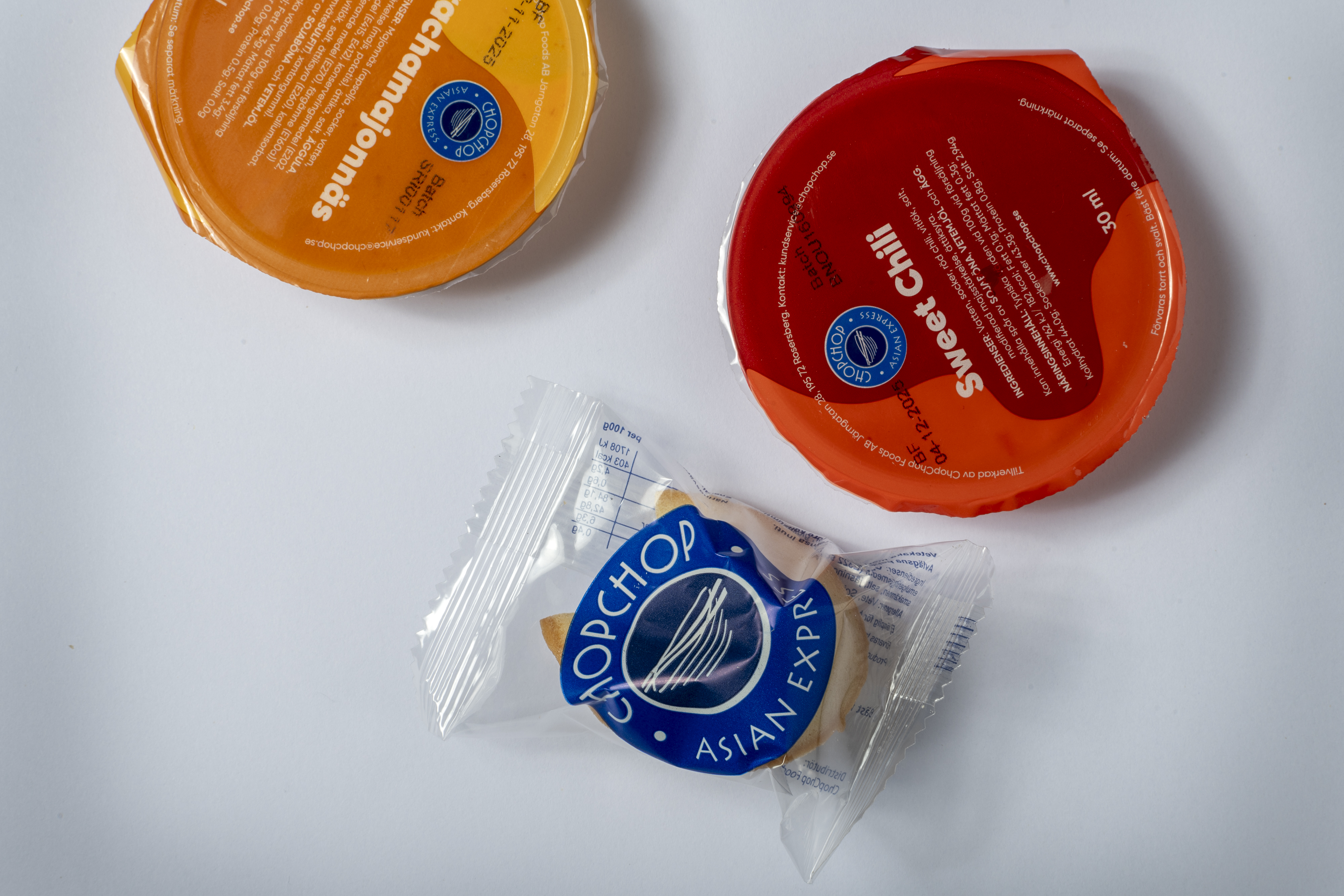 Två behållare märkta "Sriracha Orange" och "Sweet Chili" bredvid en förpackad Chop Chop Asian Express lyckokaka på vit yta.