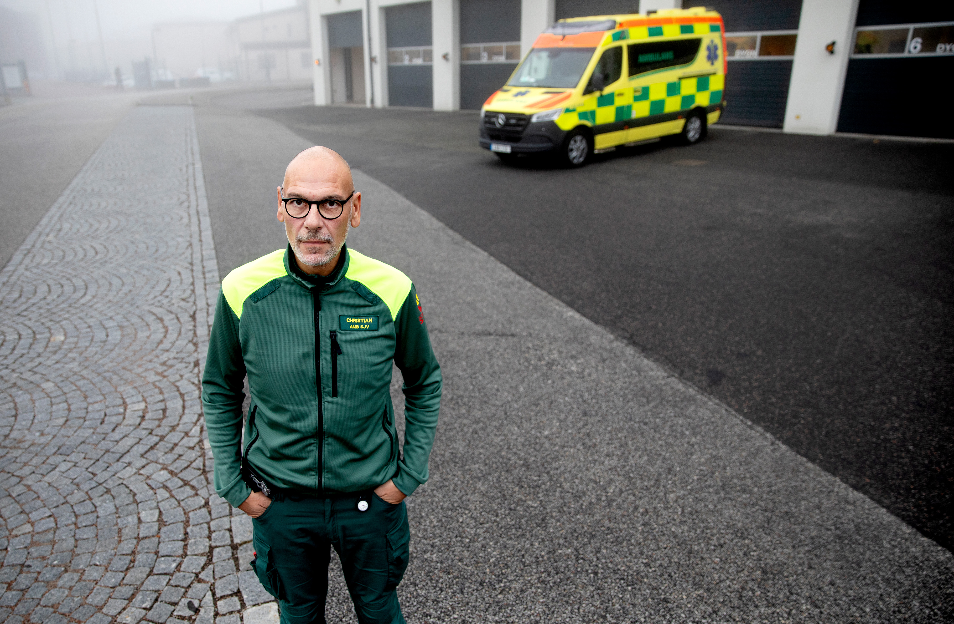 En man i grön och gul ambulansuniform står utomhus på en asfalterad yta med en ambulans och en byggnad i bakgrunden.