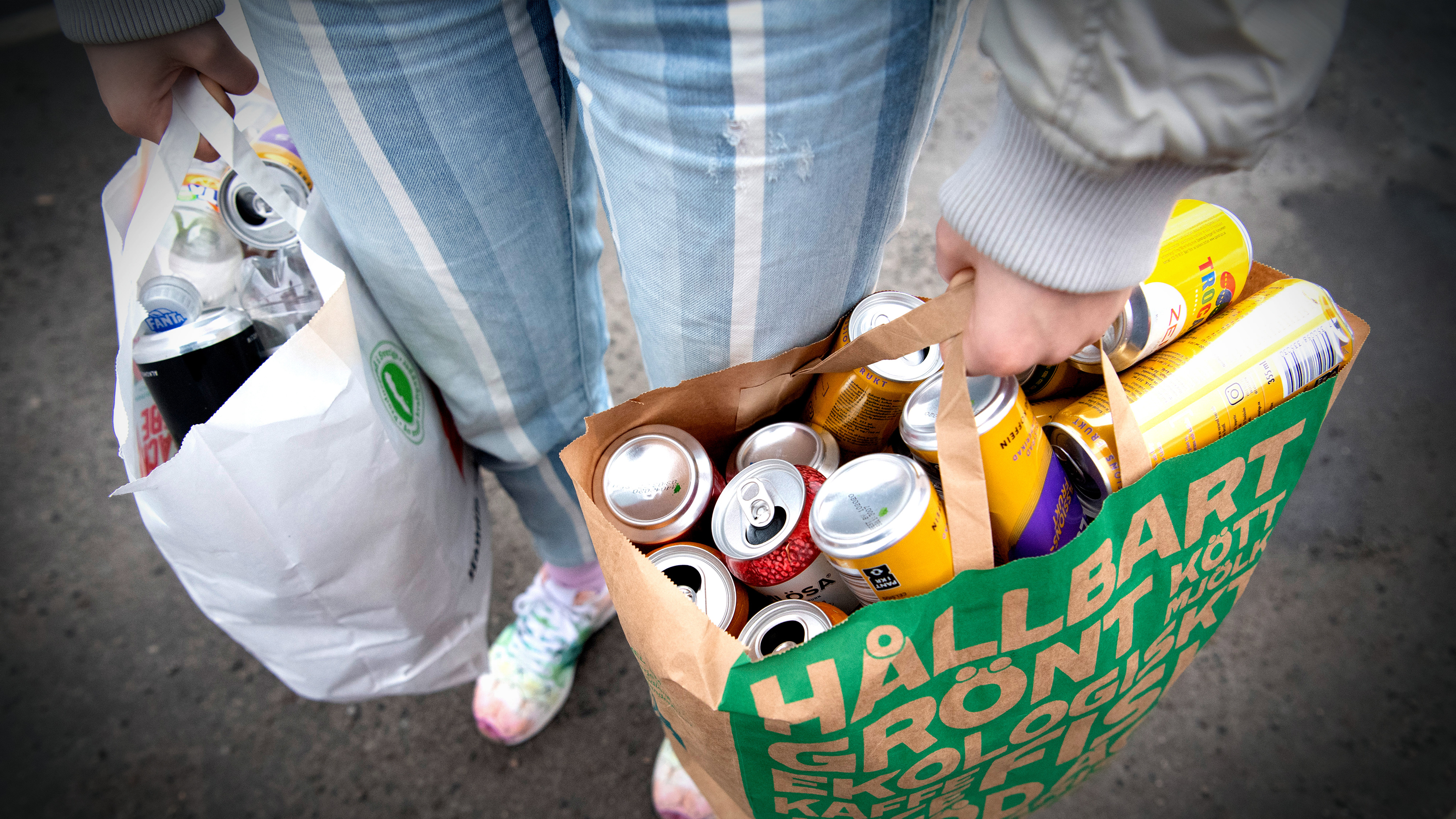 En person håller två påsar med tomma dryckesburkar och står på en stenlagd yta. Ena påsen är vit, den andra grön med svensk text.