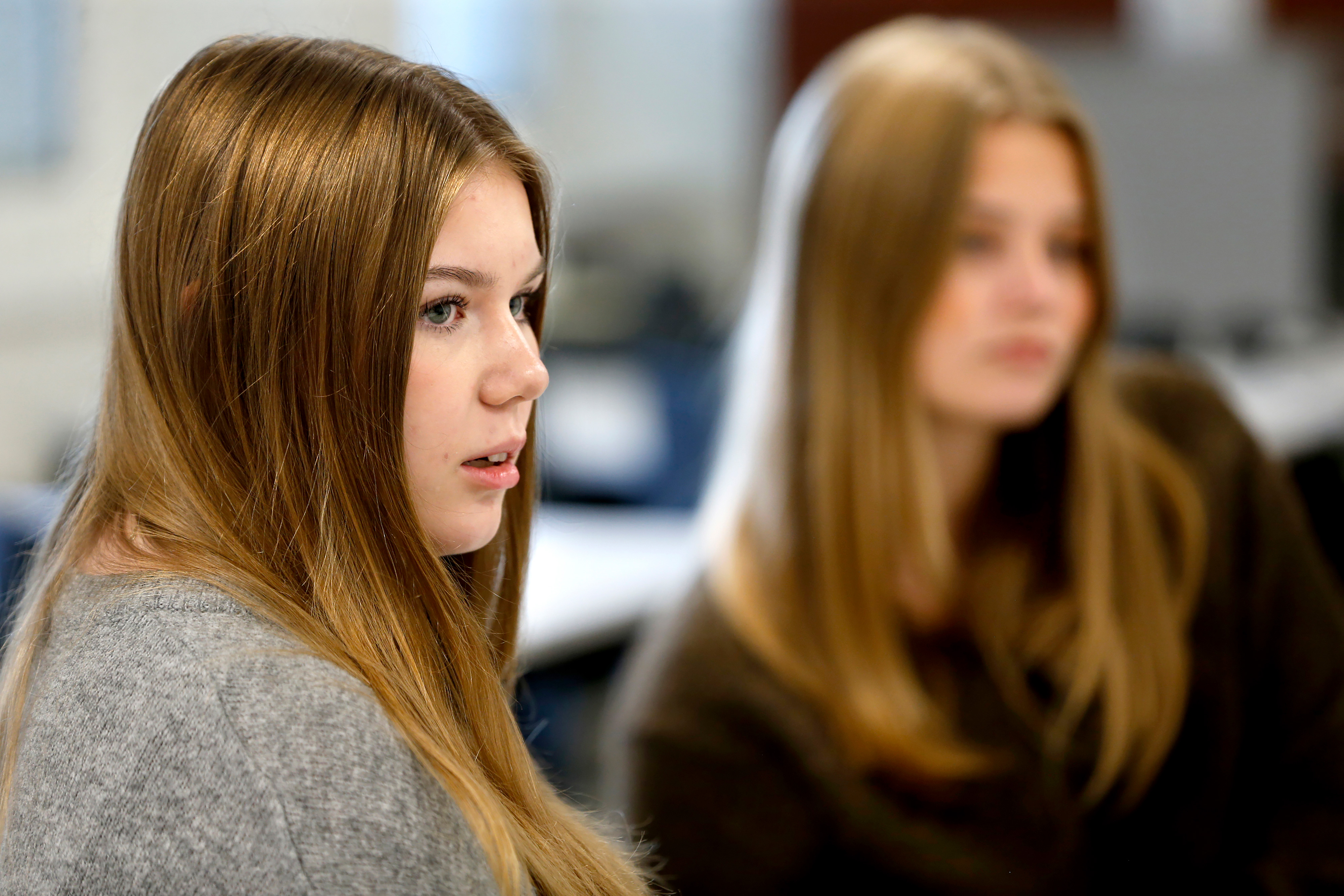 Två unga kvinnor med långt hår sitter inomhus, en tittar åt sidan och syns tydligt, den andra är i bakgrunden oskarp.