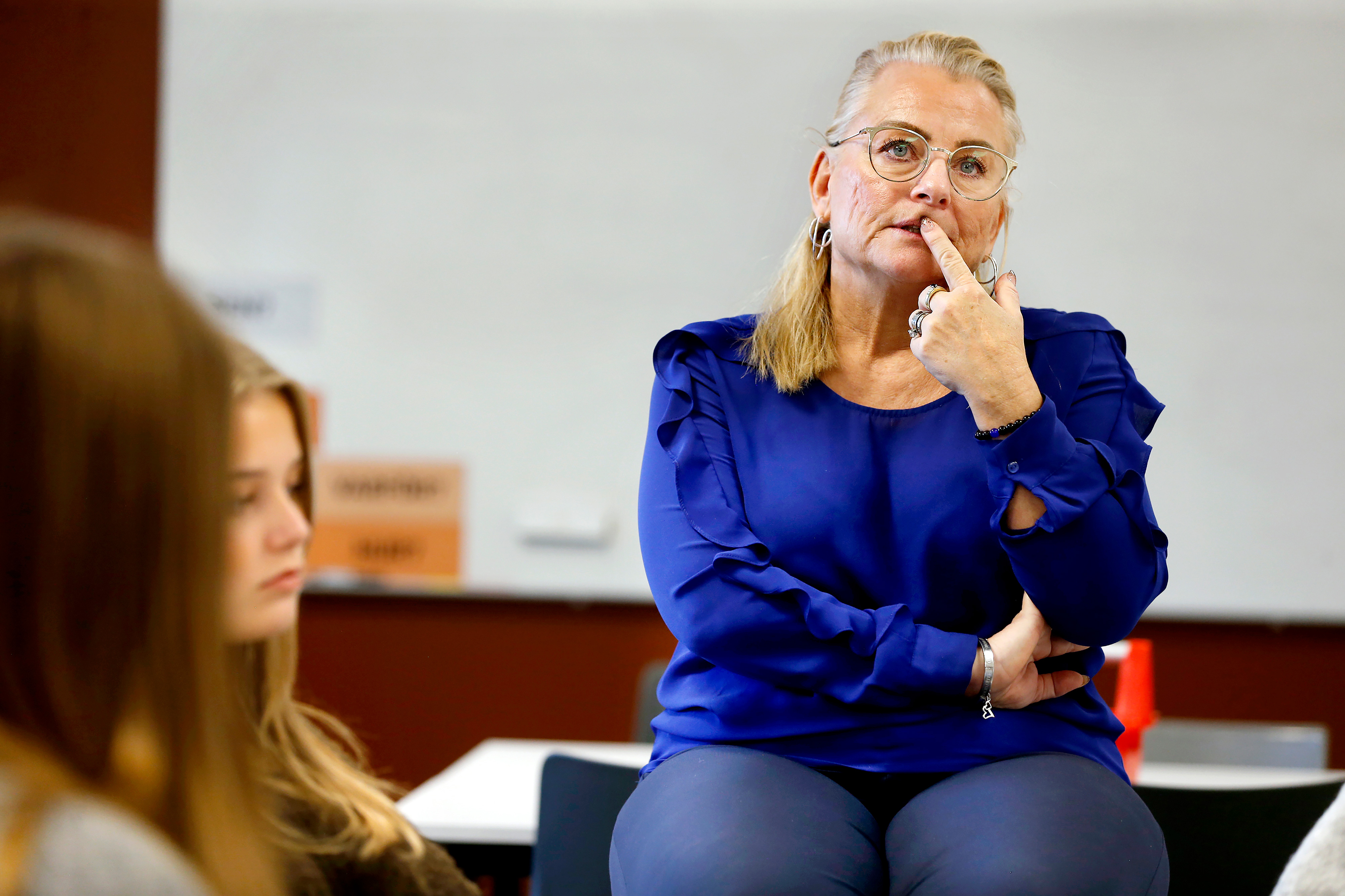 En kvinna i blå blus sitter på ett skrivbord och rör vid sitt ansikte medan en student lyssnar i förgrunden.