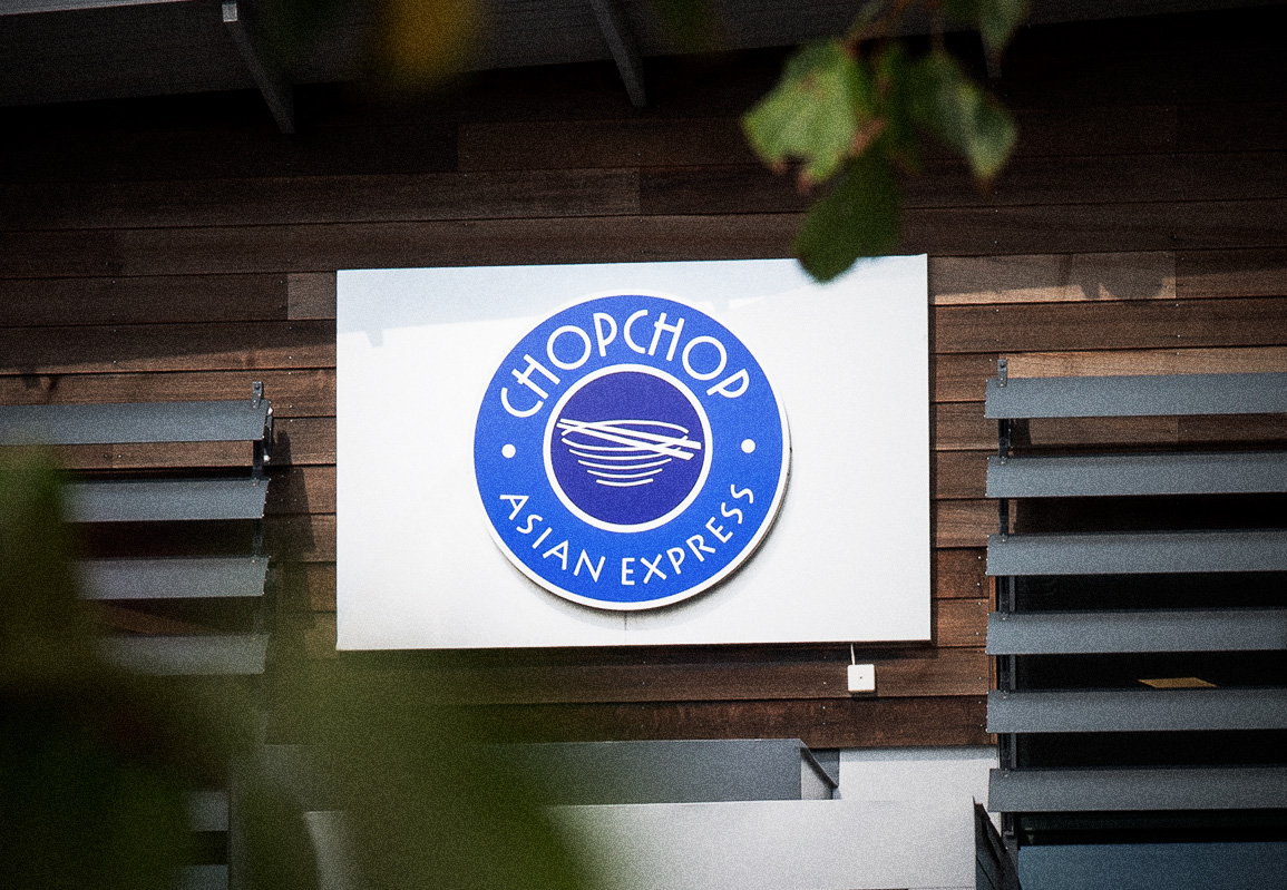En skylt med logotyp och namn "ChopChop Asian Express" är monterad på en byggnad med trä- och metallyta.