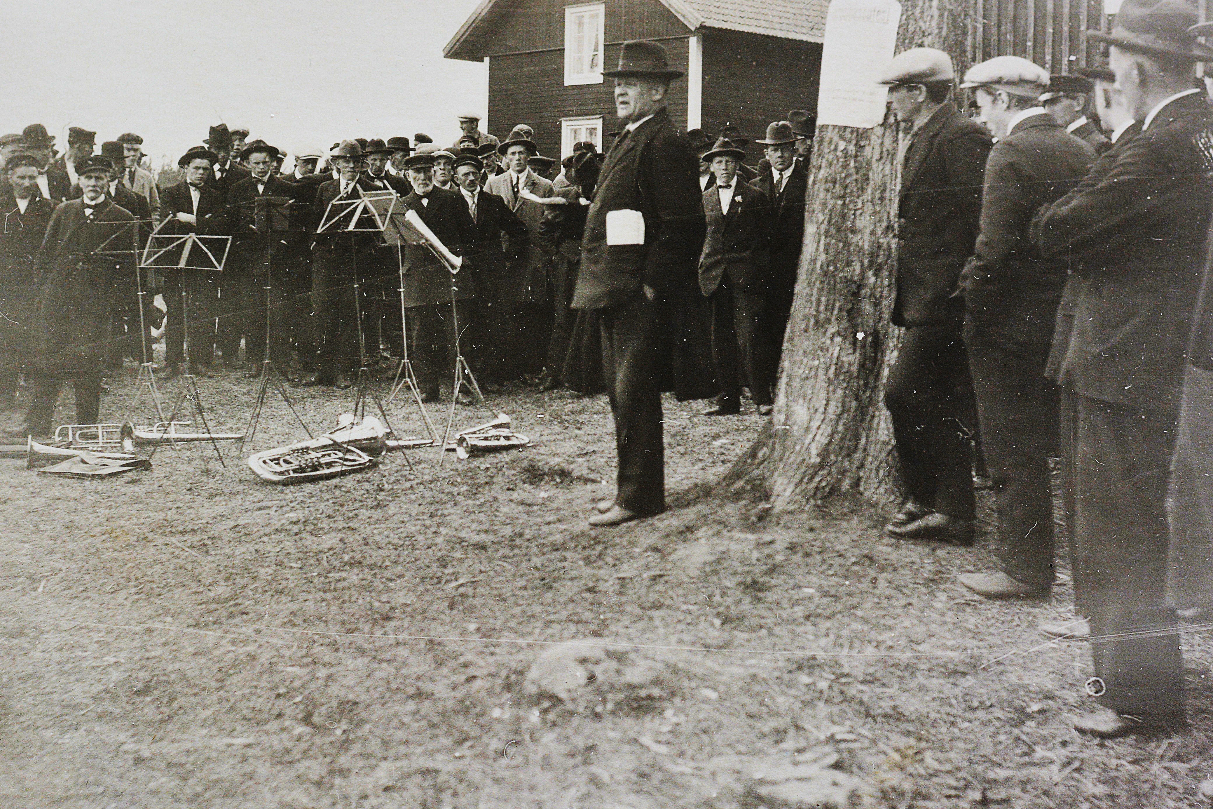 En grupp män i kostymer och hattar samlas utomhus nära ett träd medan en talare pratar; notställ och blåsinstrument ligger på marken.