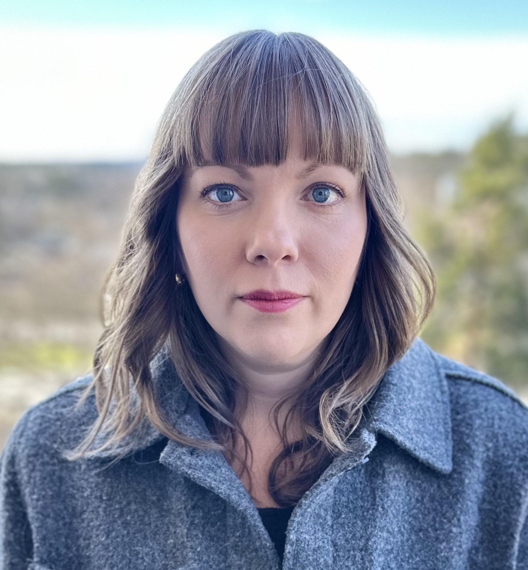Jenny Wrangborg med ljusbrunt hår och lugg står utomhus i grå kappa. I bakgrunden syns växtlighet och blå himmel.
