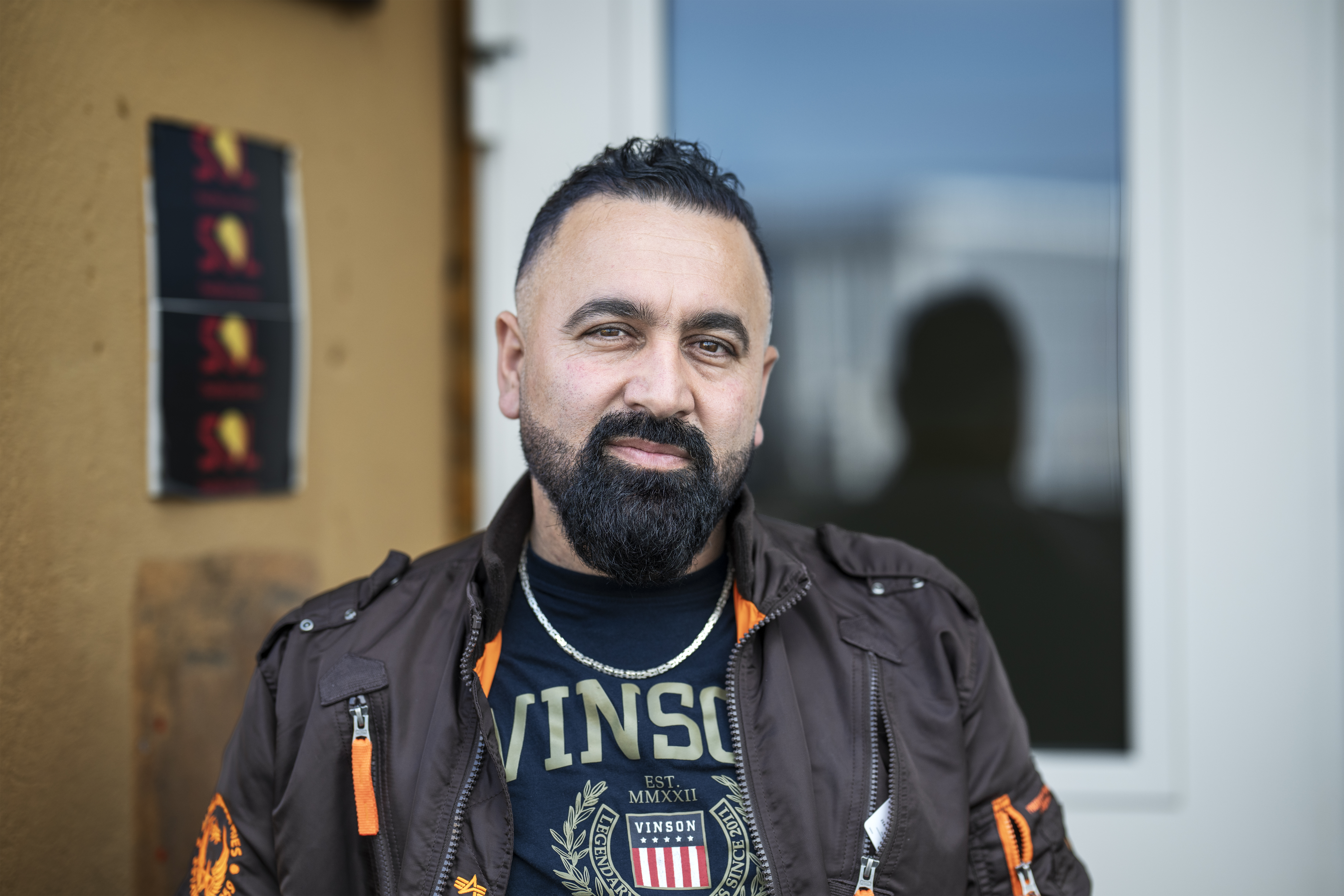 Man med skägg och kort hår har en brun jacka och står framför en byggnad.